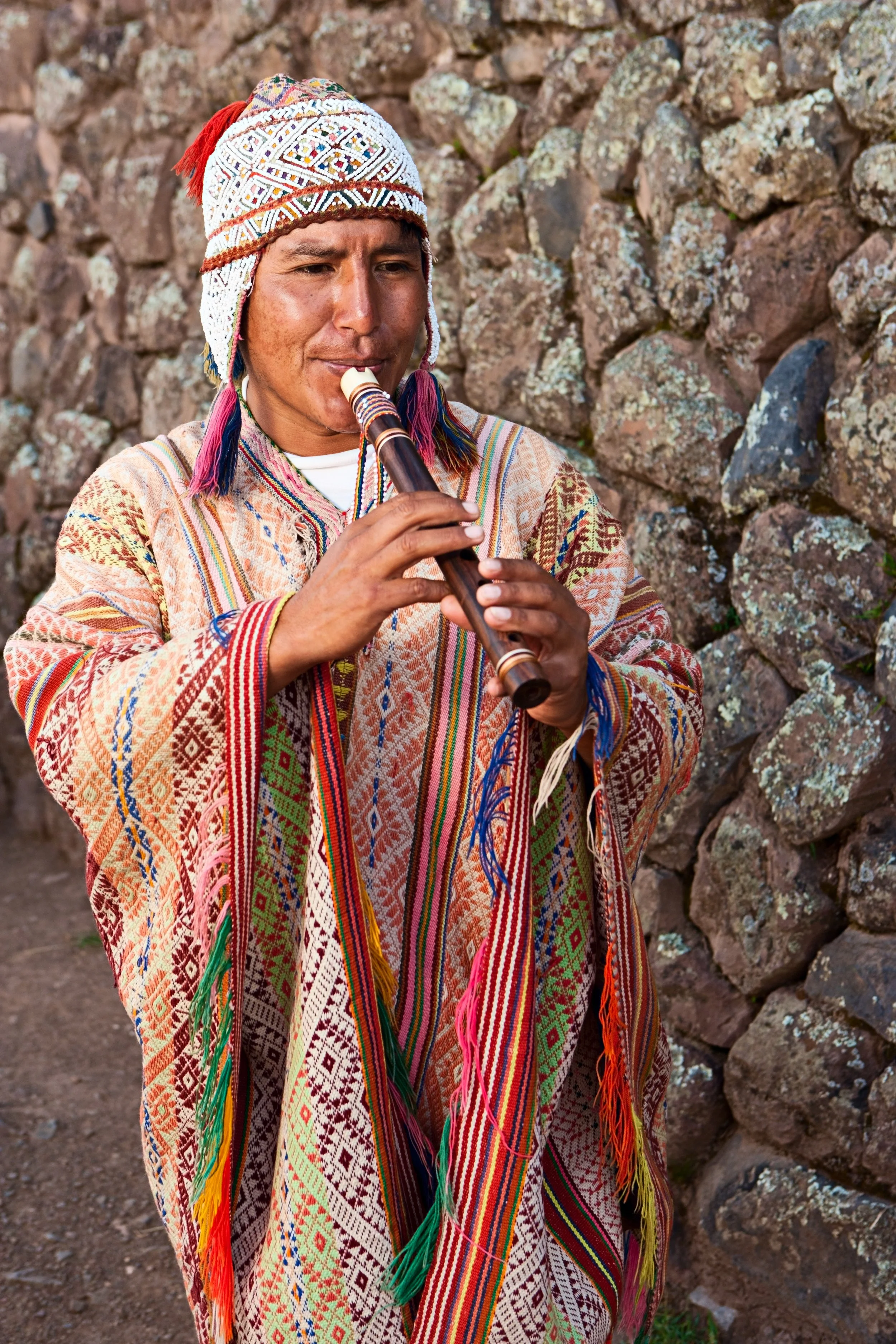 A person wearing traditional Andean clothing playing a flute outdoors near a rocky wall.