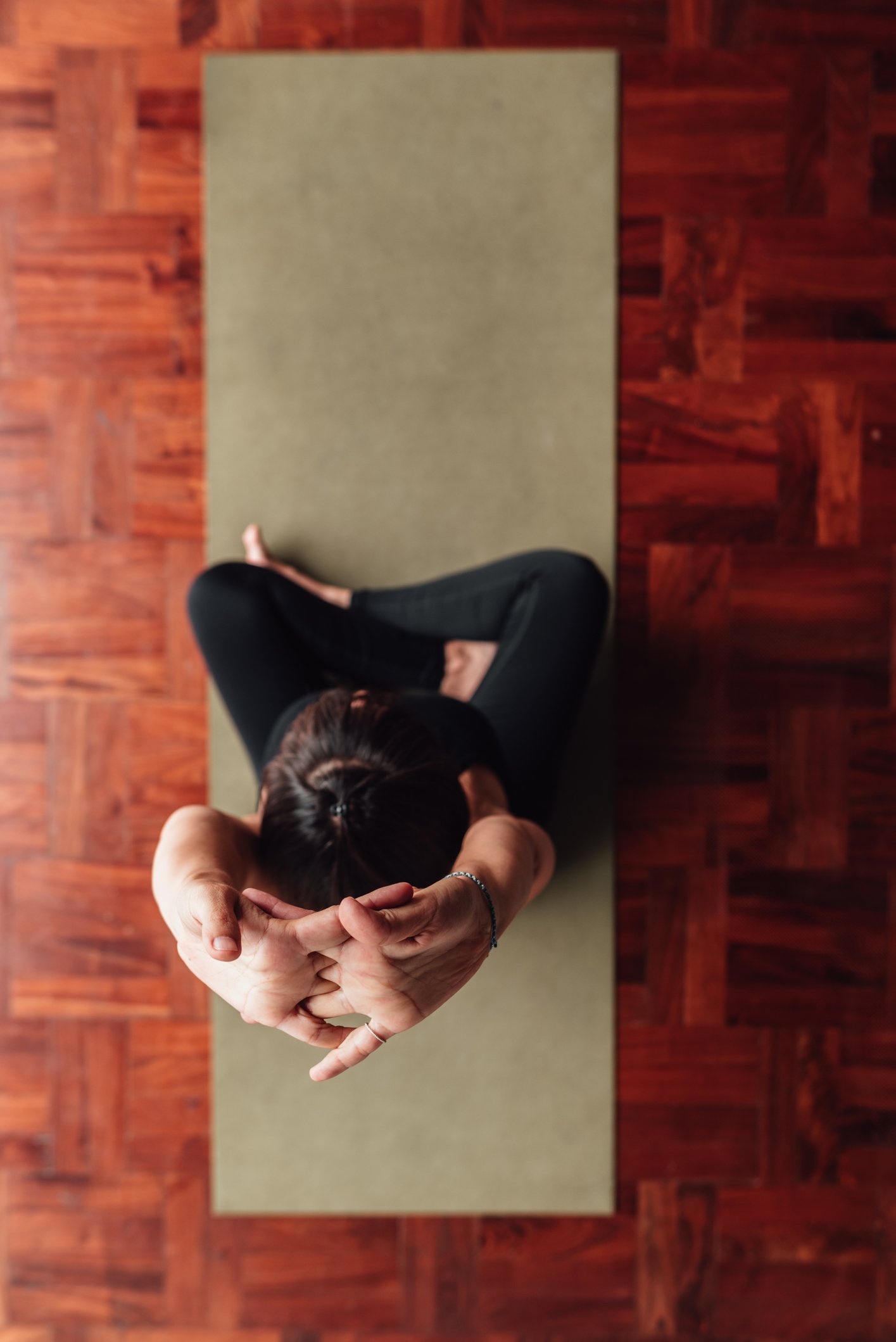 A person practicing yoga on a mat, sitting cross-legged with arms extended behind the head in a yoga pose, in a room with wooden floor.