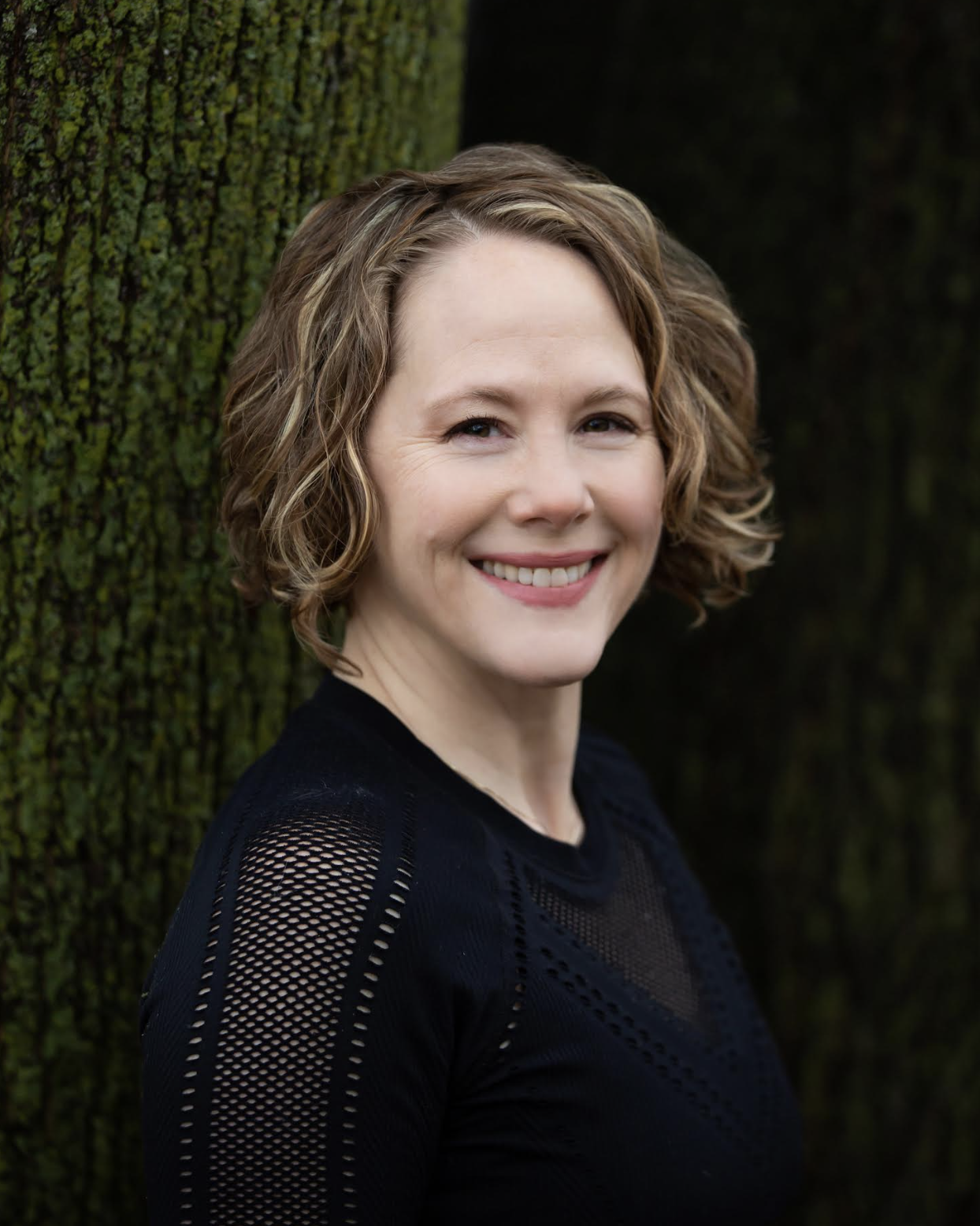 A woman with short, curly light brown hair smiling outdoors, standing next to a tree with moss-covered bark.