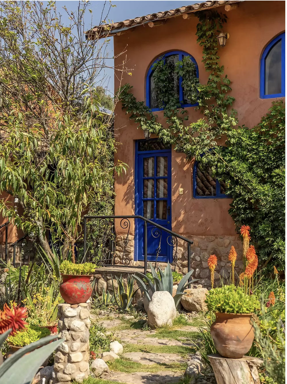 A charming orange stucco house with blue window and door frames, surrounded by lush green plants, climbing vines, and a vibrant garden with colorful flowers in terracotta pots.