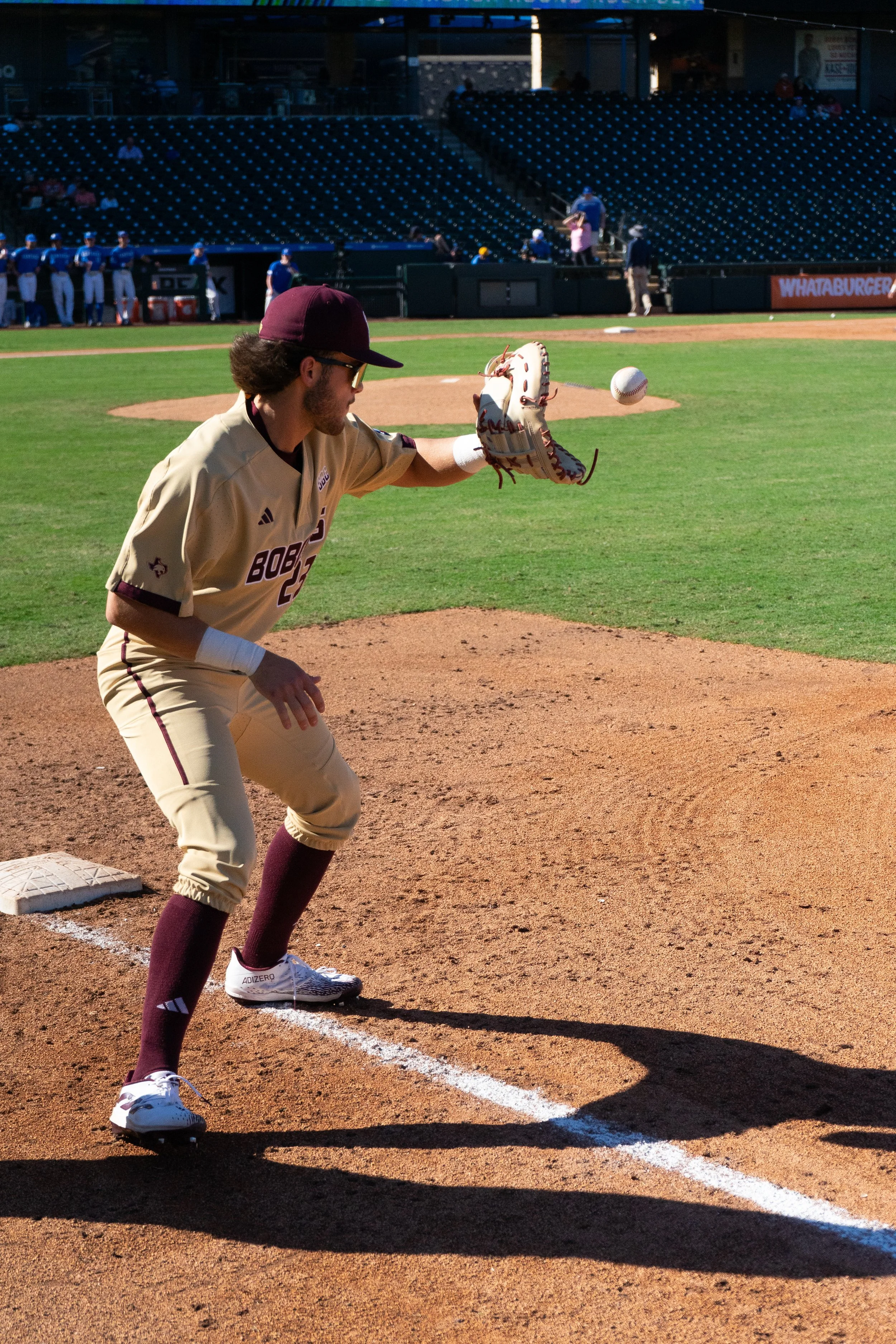 A baseball player in uniform practicing fielding skills on a baseball field, with the ball mid-air near his glove and the pitcher’s mound and bleachers in the background.