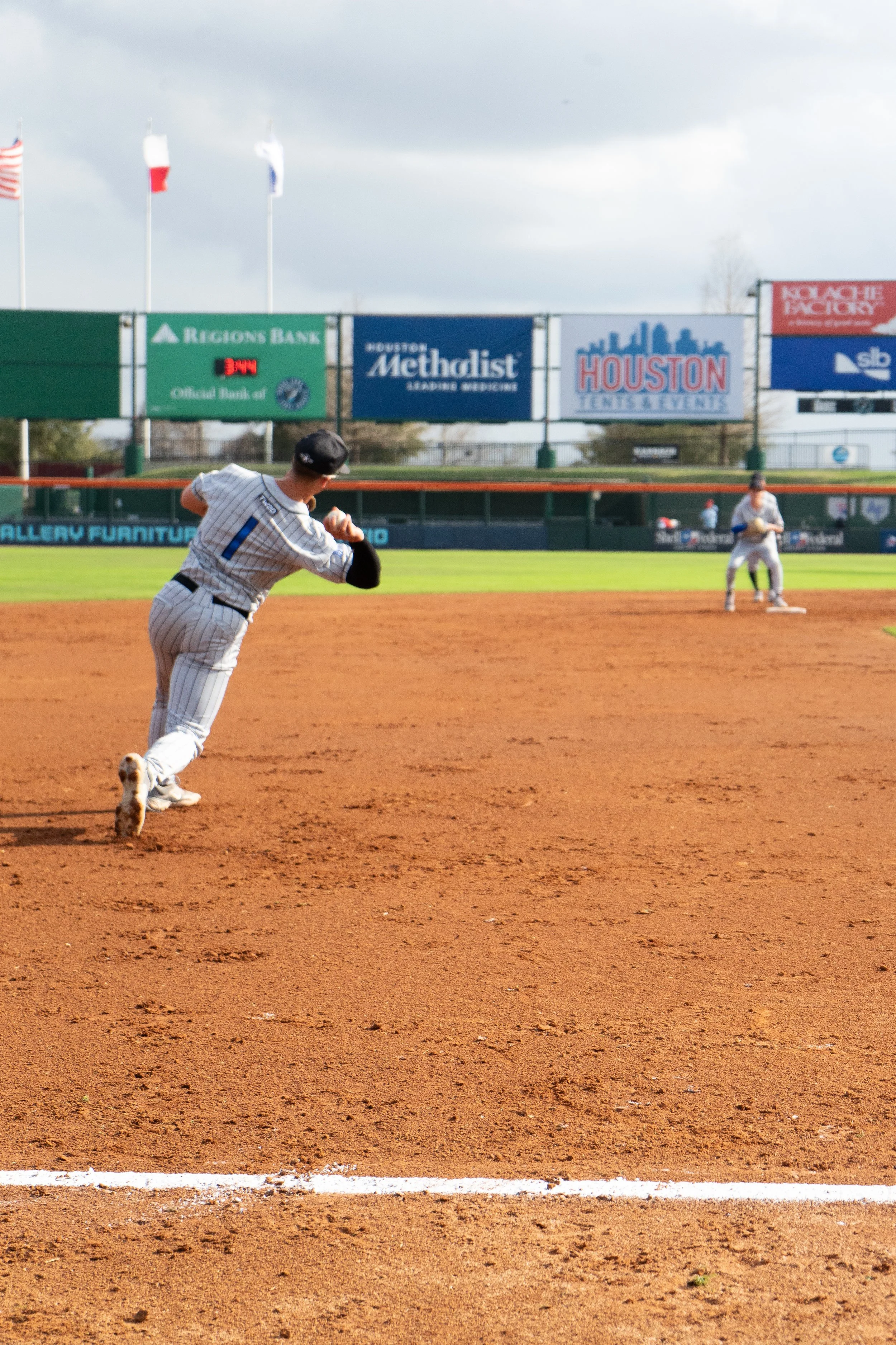 A baseball player in a pinstripe uniform running on the field with another player positioned behind in the outfield, at a baseball stadium with advertisements and flags in the background.
