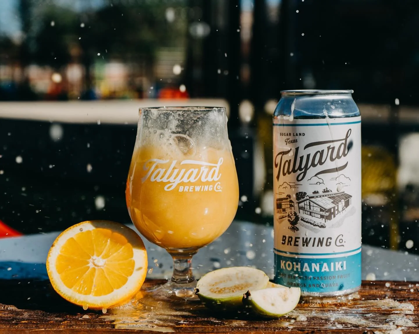 A can of Talyard Brewing Kohanaiki beer, a filled beer glass with the same logo, half an orange, and slices of lime on a wet wooden surface with splashes of water, outdoors with a blurred background.