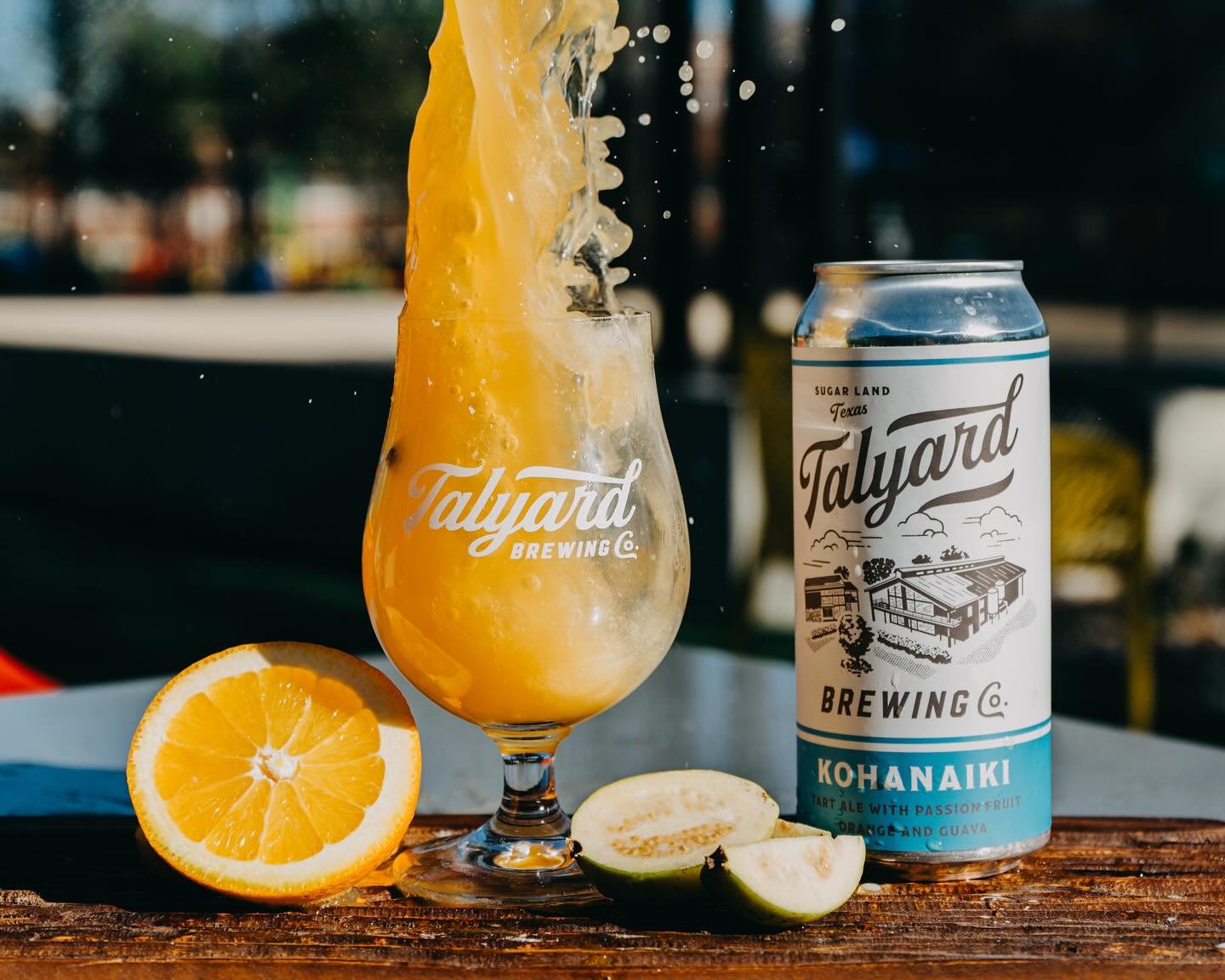 A can of Talyard Brewing Co. Kahanaiki craft beer with passion fruit, orange, and guava flavor, placed on a wooden surface. In front, a sliced orange, cucumber, and lemon are displayed. A filled glass with the same beer is behind the ingredients, with the beer being poured into the glass.