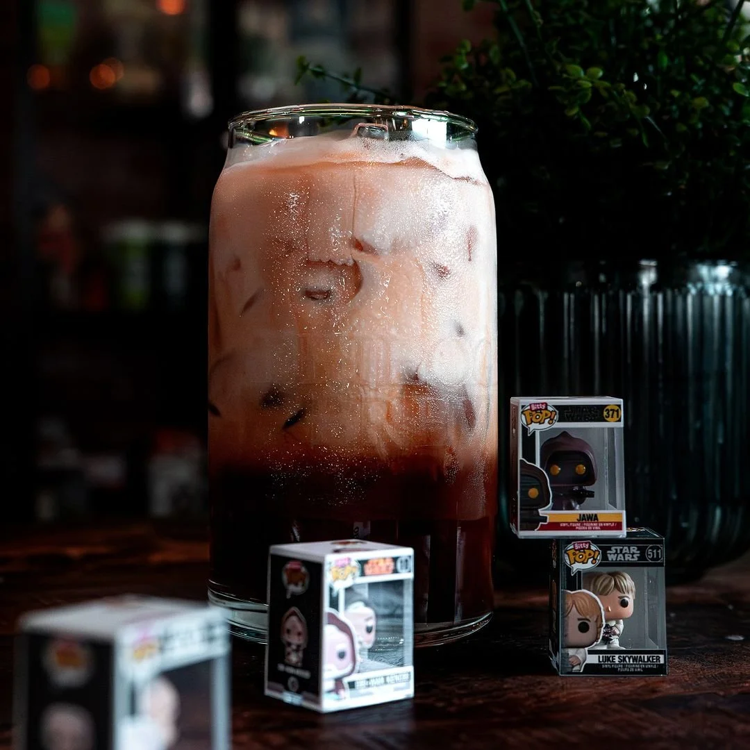 A glass of iced coffee with milk and ice cubes on a wooden surface, surrounded by three Funko Pop! figures of Star Wars characters, Jowa, Luke Skywalker, and another one partially visible.