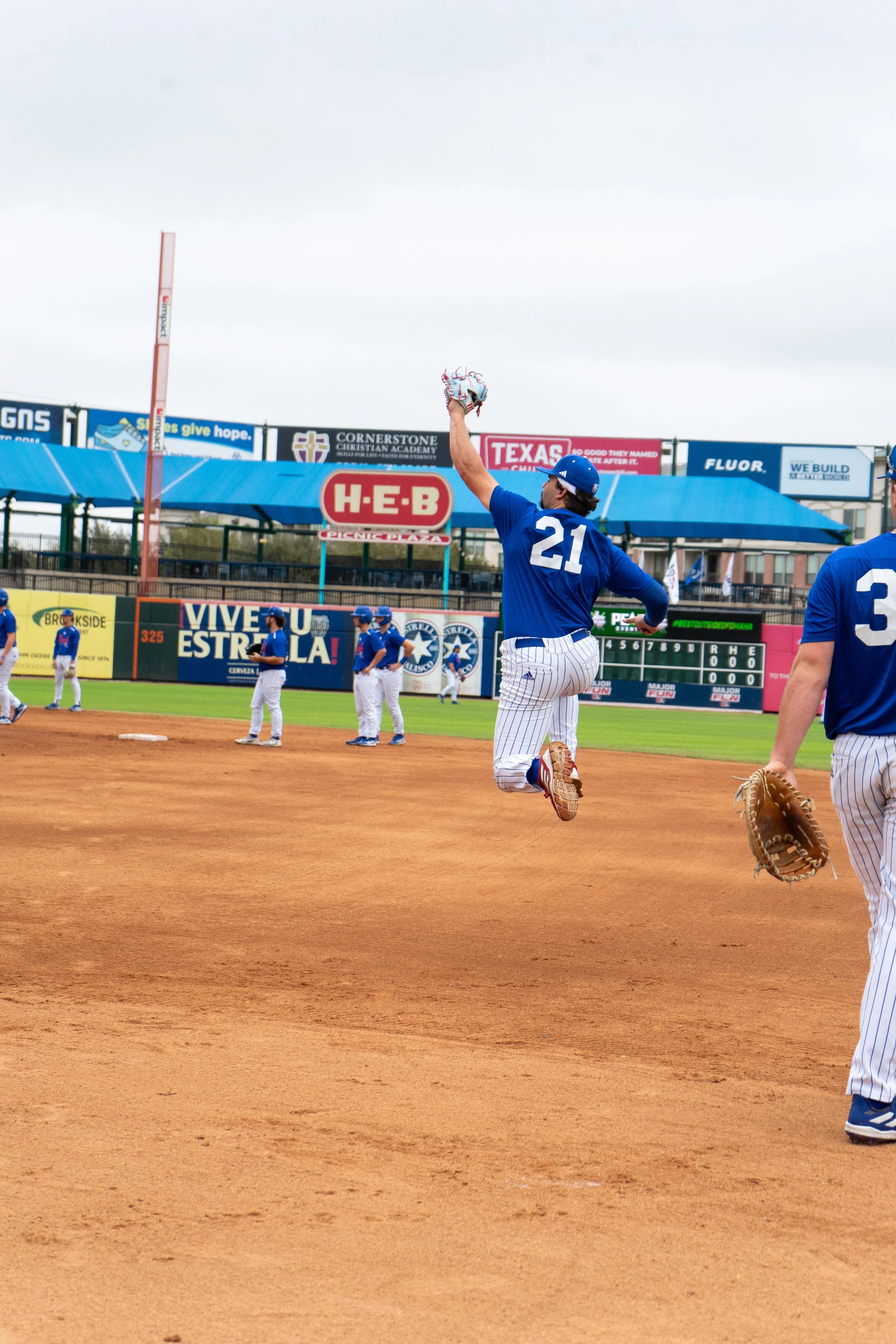 Baseball players on the field during a game, with one player jumping and reaching up to catch a ball, wearing a blue jersey with the number 21.