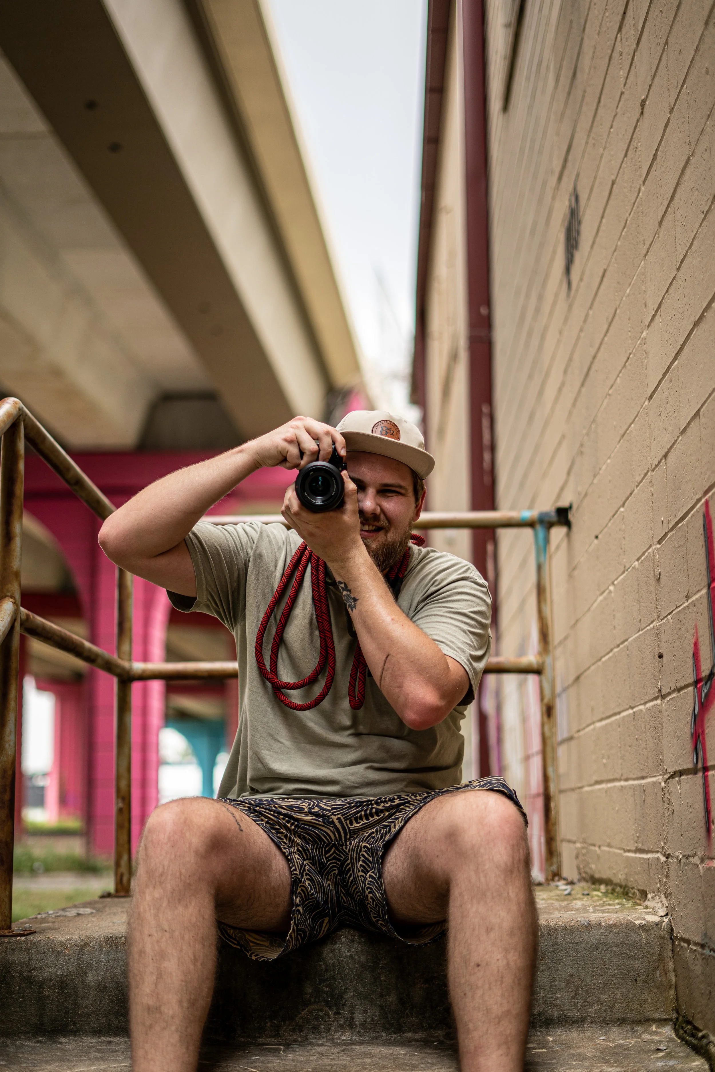 A man sitting on concrete steps, taking a photo with a camera. He's wearing a tan cap, a beige t-shirt, and patterned shorts, with a red rope around his neck. He is smiling and looking into the camera lens.