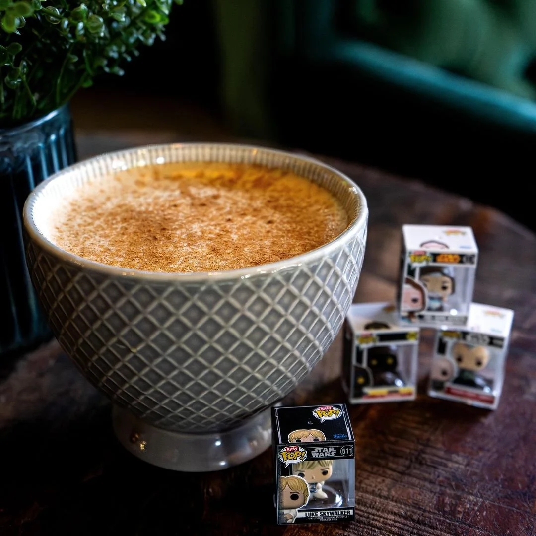 Large decorative cup filled with a frothy beverage, positioned on a wooden table next to a potted plant and several Funko Pop! Star Wars Luke Skywalker figures.