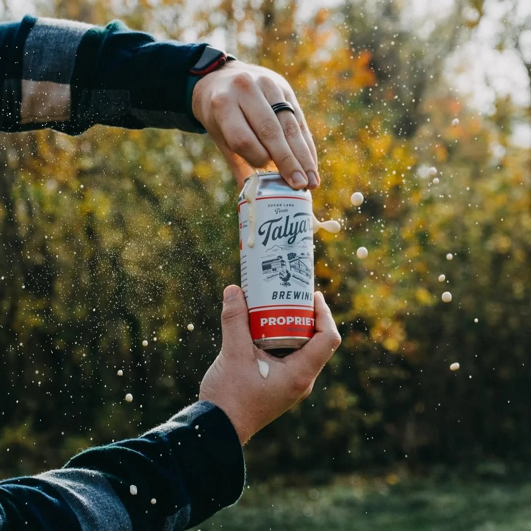 Two people opening a can of beer outdoors, with foam splashing out. One person's hand is visible, holding the can, while the other is pulling the tab open. The background features blurred autumn trees.