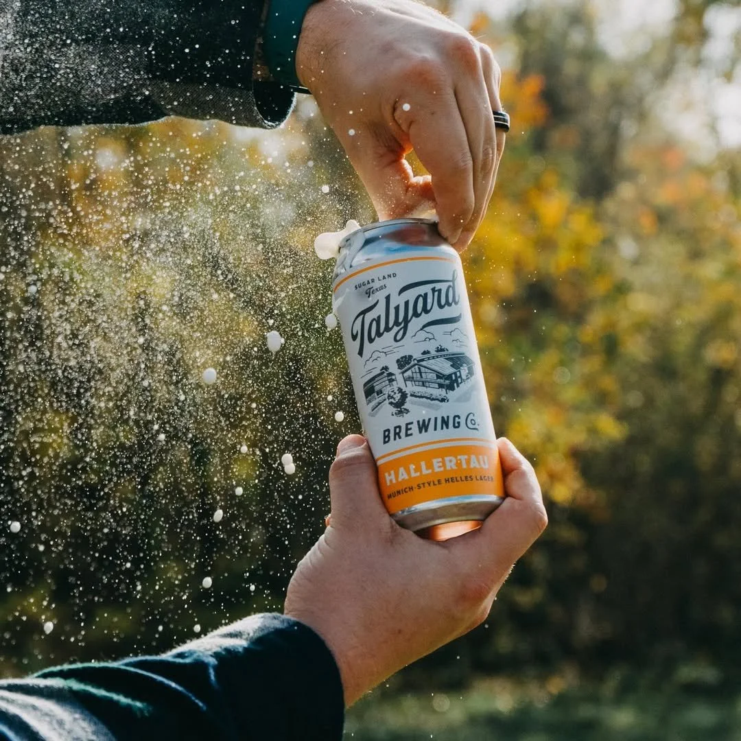 Person opening a can of Talyard Brewing's Hallertau beer outdoors with a blurred background of autumn trees.