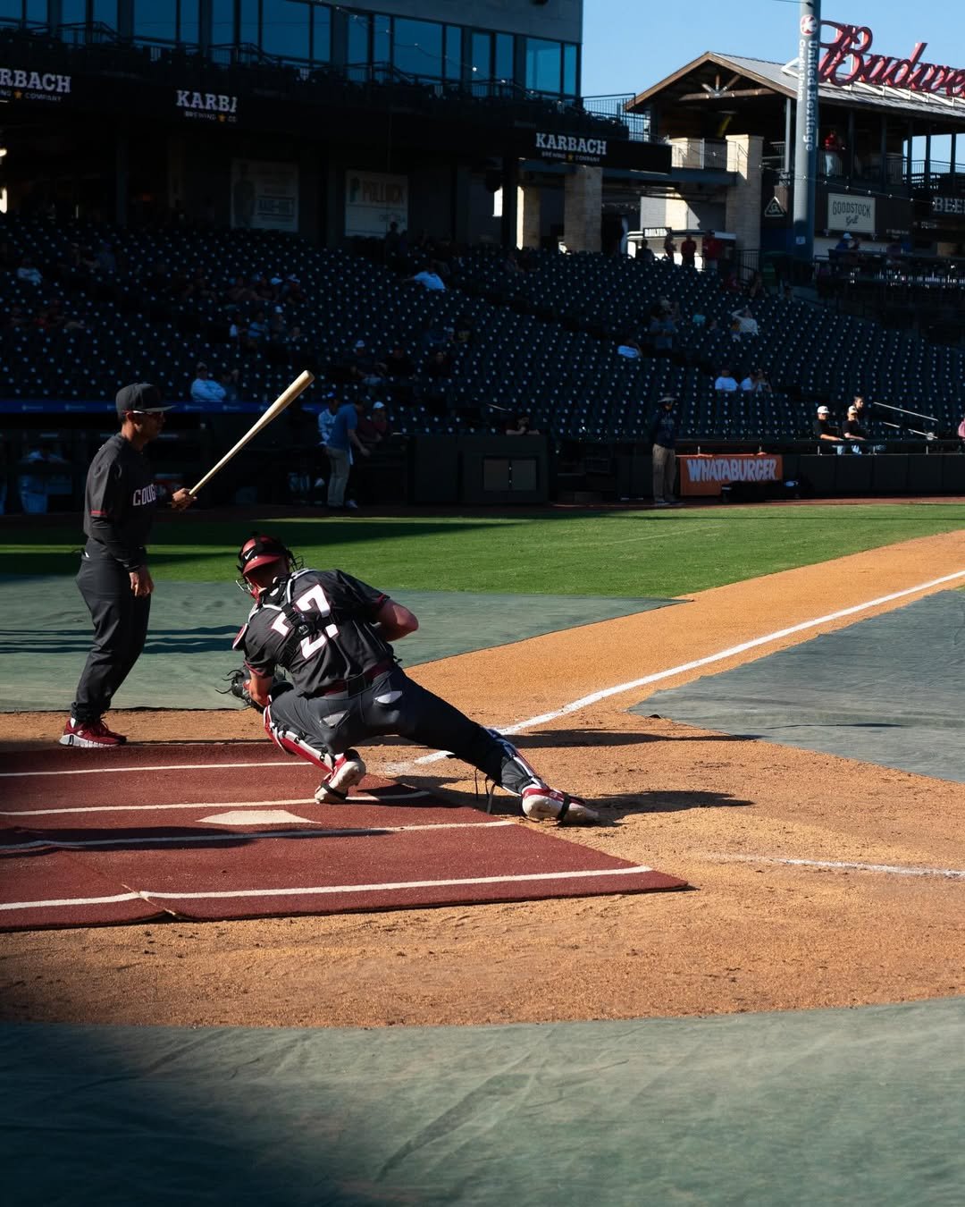A baseball player at bat, a catcher crouched behind home plate, and an umpire standing nearby on a baseball field in a stadium with empty seats and advertisements visible in the background.