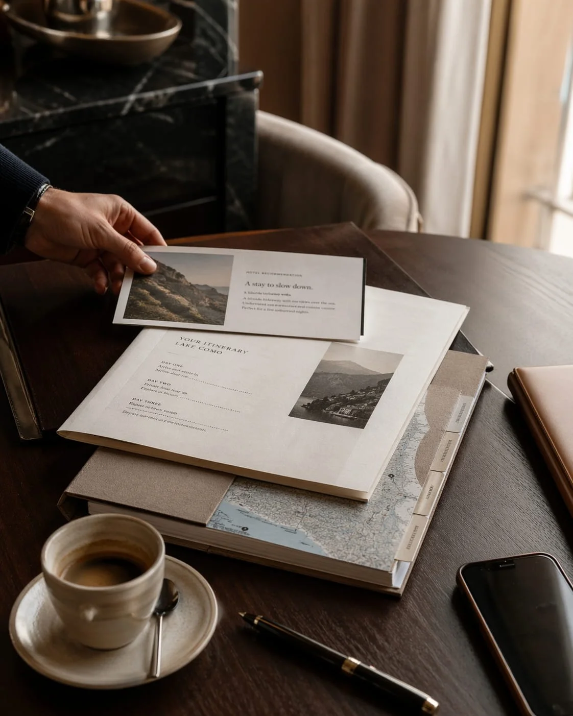 Hotel recommendation card, itinerary packet, map tabs, coffee, and phone arranged on a dark wood table.