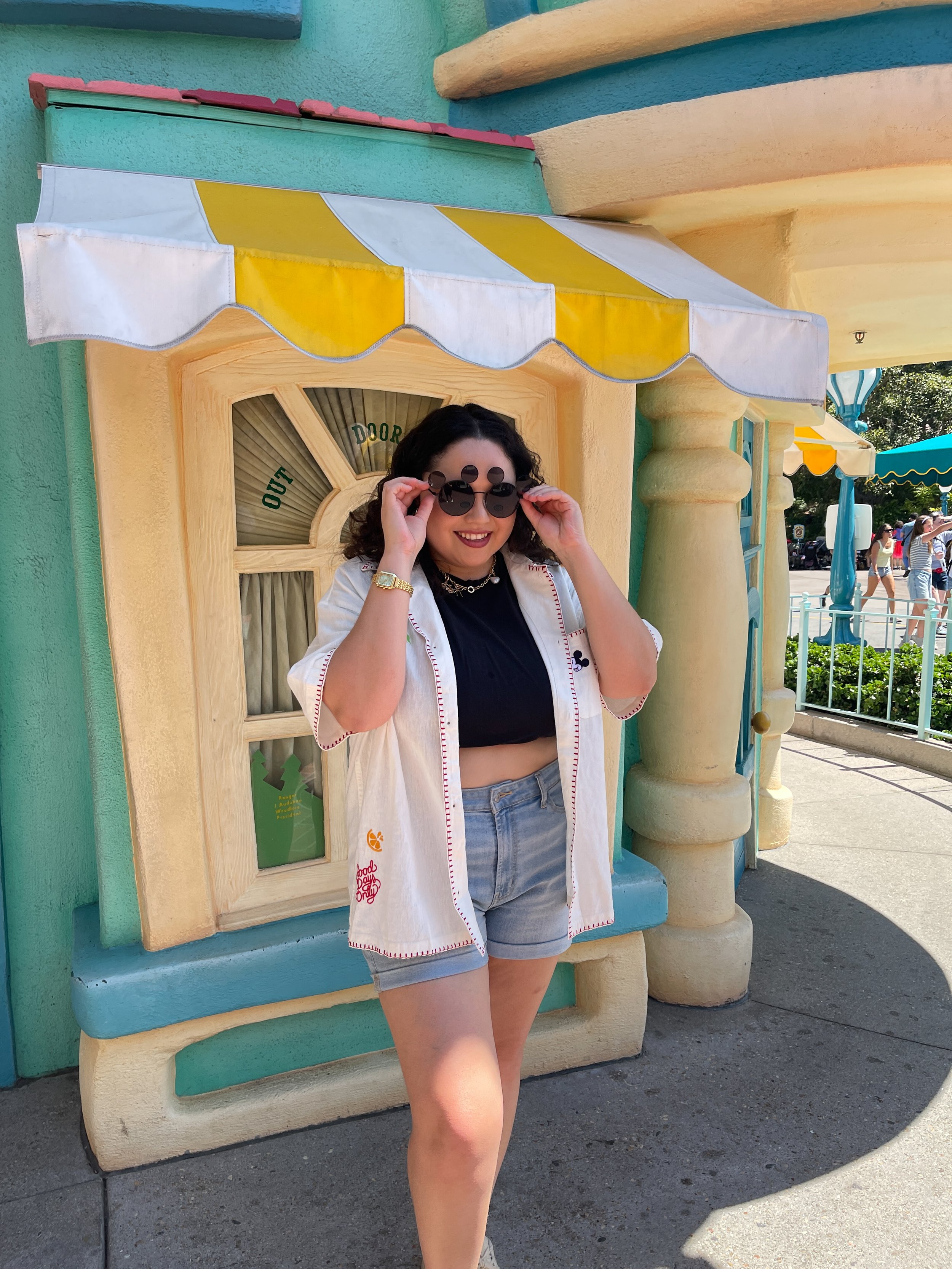 Woman with curly dark hair wearing sunglasses, a black crop top, a short-sleeved white open shirt with red stitching, and light denim shorts standing outside a colorful cartoon-style building with pastel yellow, teal, and pink walls, and a yellow and white striped awning.