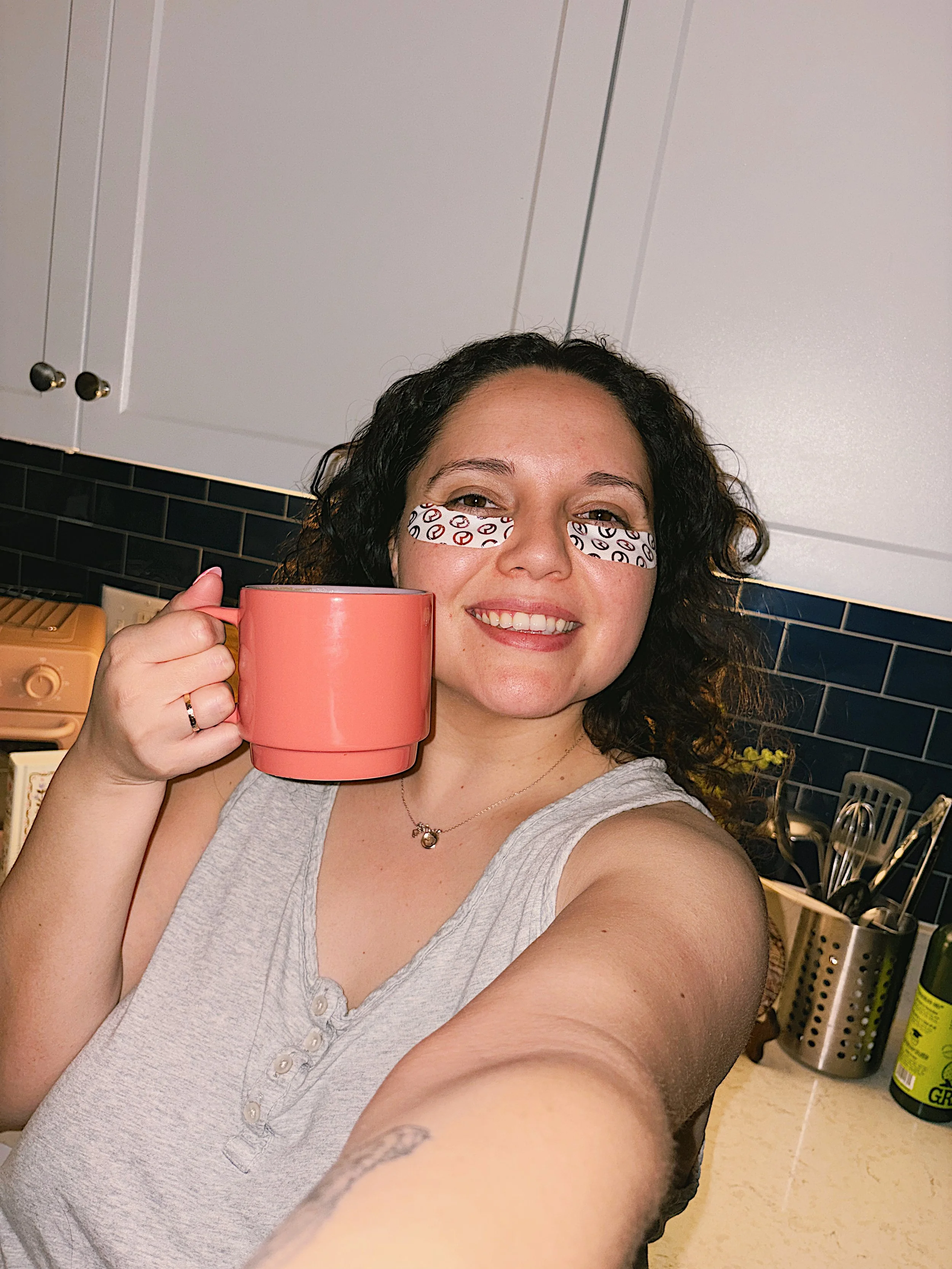 A woman smiling in a kitchen, holding a pink mug, with eye black sporting a black and white swirl pattern on her face.