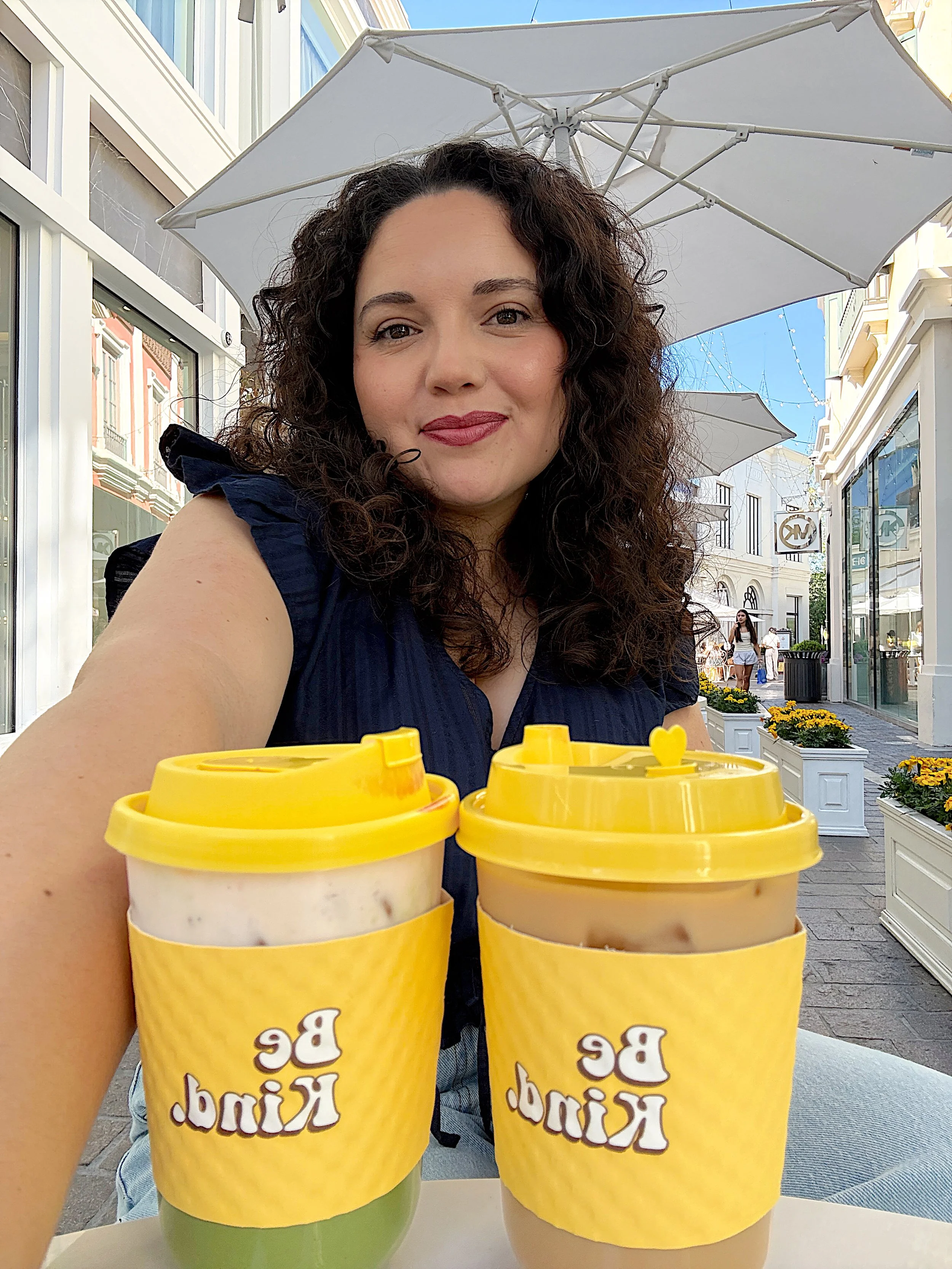 Woman taking selfie outdoors at a shopping or dining area with two cups of iced drinks in front of her, sunlight, storefronts, and pedestrians in the background.