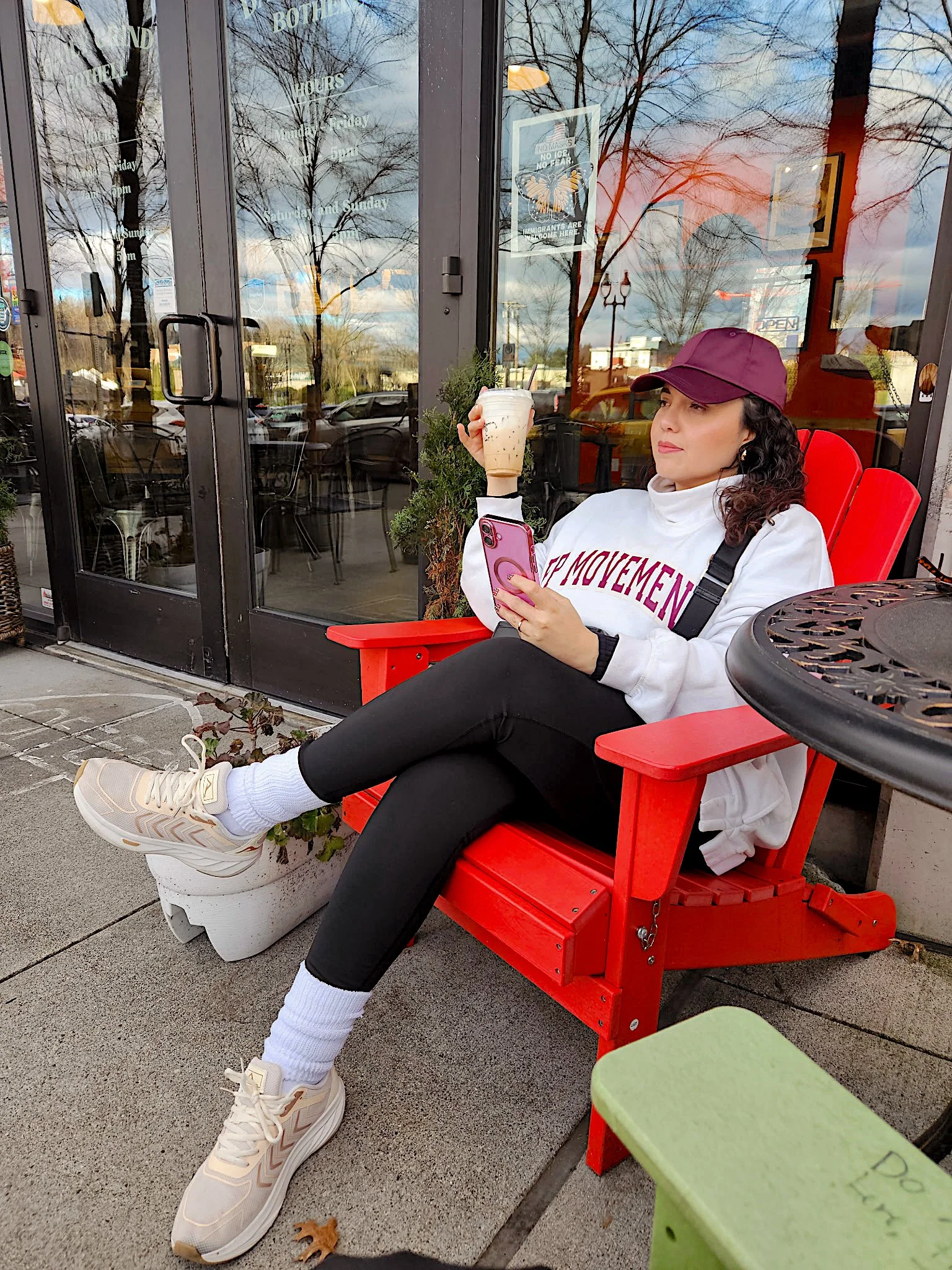 A woman sitting outside a cafe on a red chair, holding a pink phone in one hand and a coffee drink in the other, dressed in a white sweatshirt, black pants, cream sneakers, white socks, and a purple cap, with a leaf on the ground near her feet and tree reflections on the cafe glass door.
