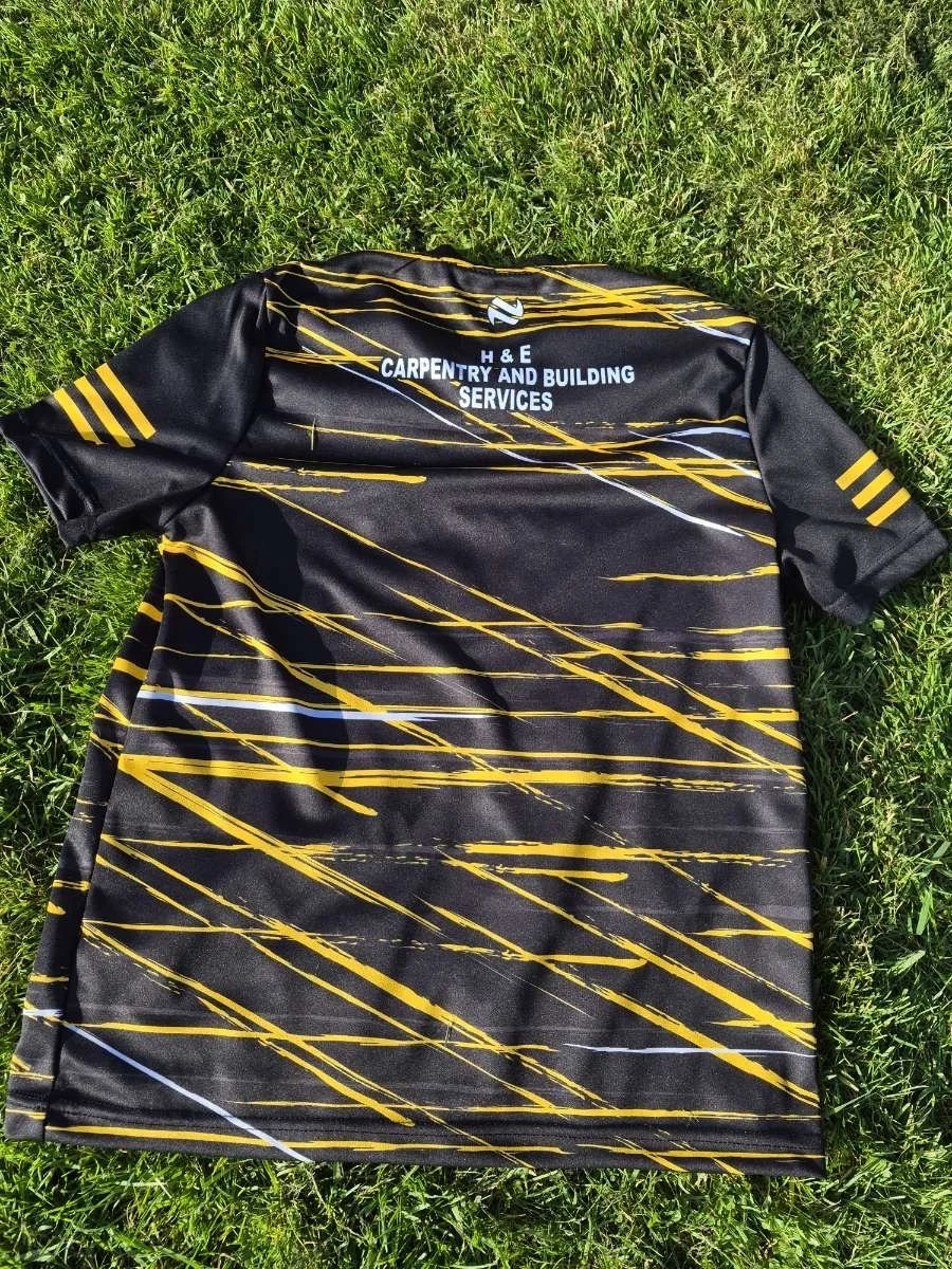 A black sports jersey with yellow and white streaks is lying on green grass. The jersey features the logo and text for 'H & E Carpentry and Building Services' on the back, along with a small white logo near the top.