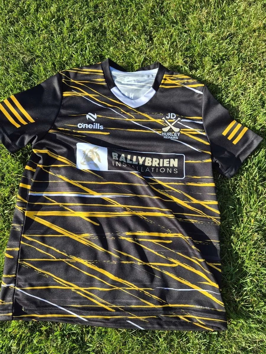 Black sports jersey with yellow and white streaks lying on green grass, featuring various logos, including 'JD Hurley Repairs' and 'Ballybrien Installations.'