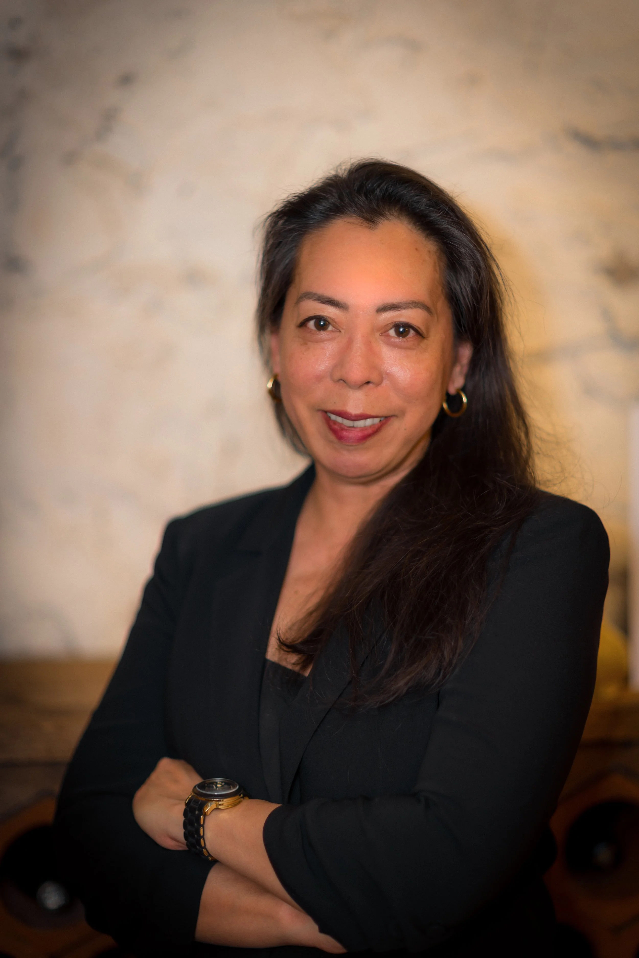 A woman with long black hair, wearing gold hoop earrings and a black blazer, stands with her arms crossed and smiles at the camera in front of a beige textured wall.