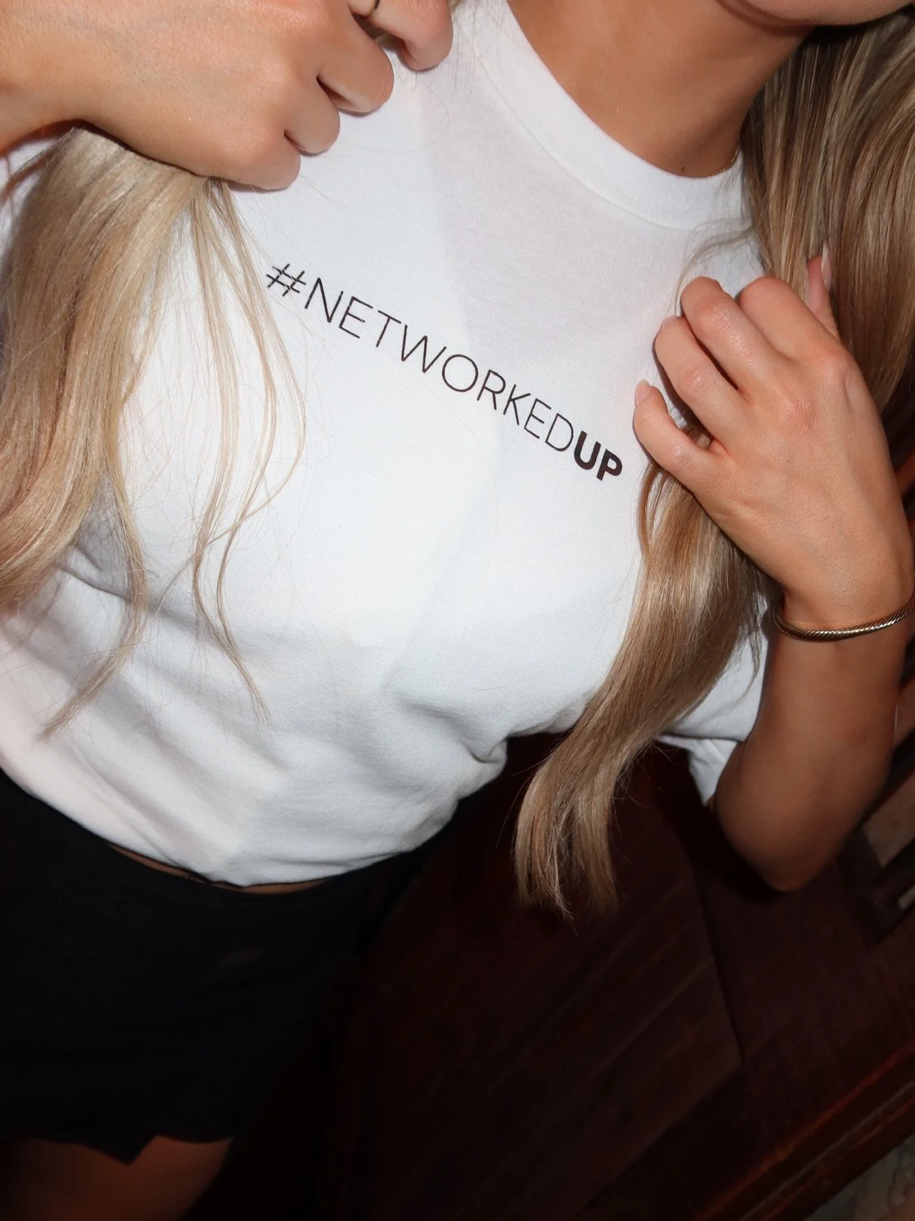 Close-up of a woman wearing a white t-shirt with the hashtag '#NETWORKEDUP' printed on it, and her hand resting on her shoulder.
