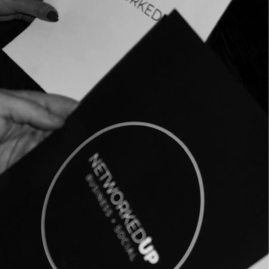 Close-up of a person's hand holding a black card or booklet with the text 'NETWORKED UP' and a website URL.