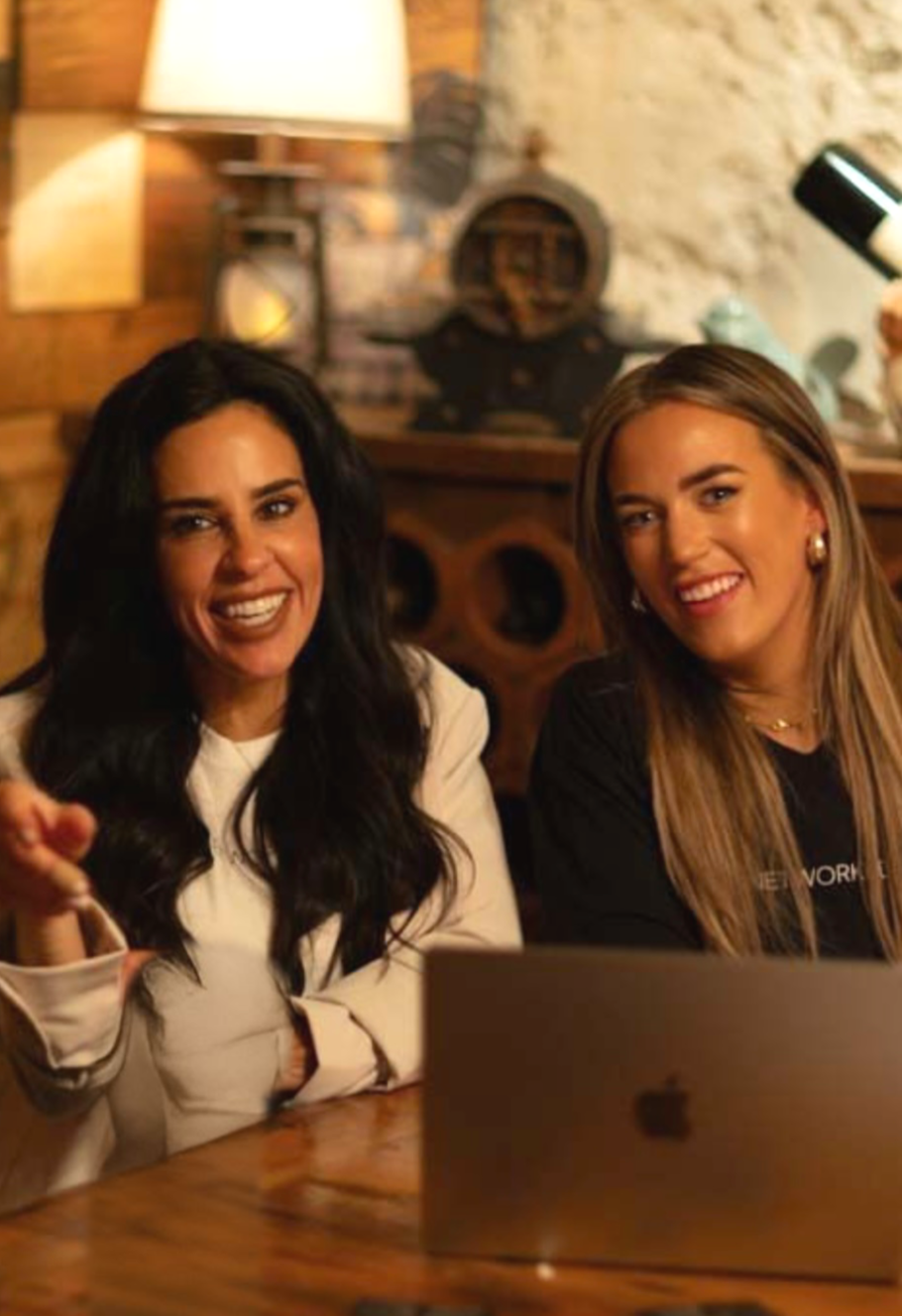 Two women sitting at a wooden table, smiling at the camera, with a laptop in front of them, in a cozy, warmly lit room.