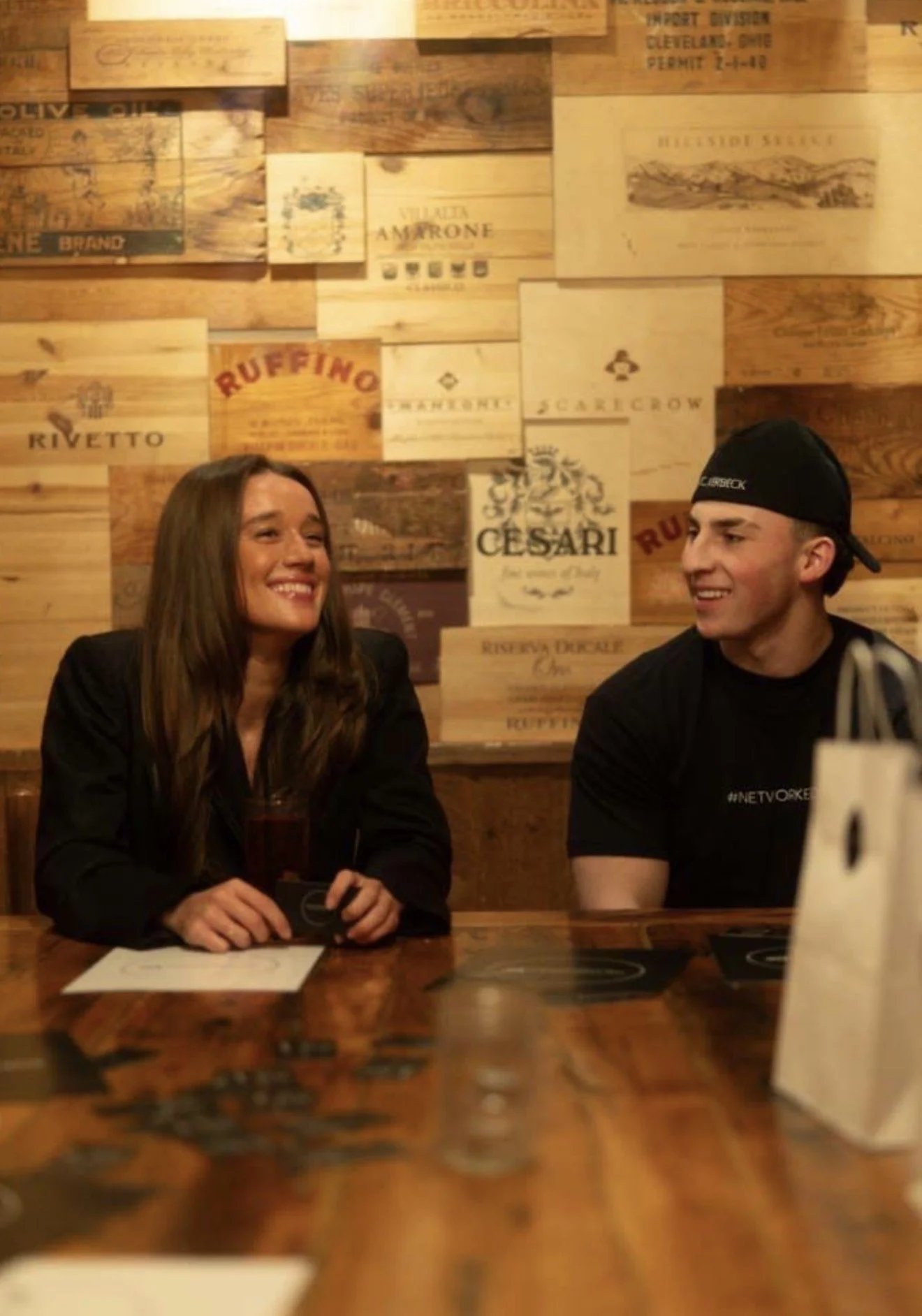 A woman and a man sit at a wooden table in a restaurant or bar, smiling and engaging in conversation. The woman has long brown hair and is wearing a black blazer, while the man is wearing a black t-shirt and a backward baseball cap. The background features a wall made of wooden panels with various vintage-style signs and logos.