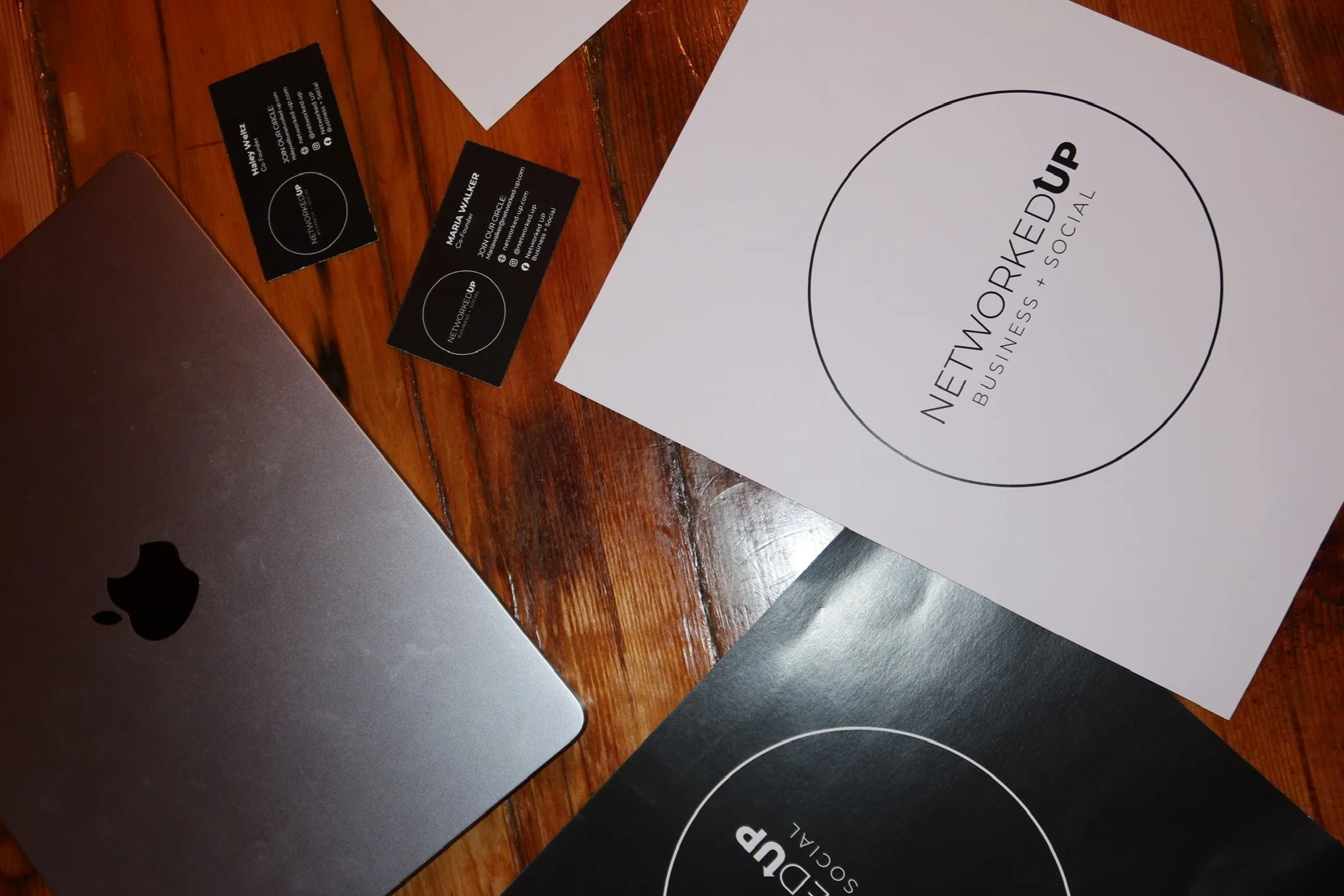 Business cards, a laptop, and printed materials labeled "NETWORKED UP BUSINESS + SOCIAL" on a wooden table.