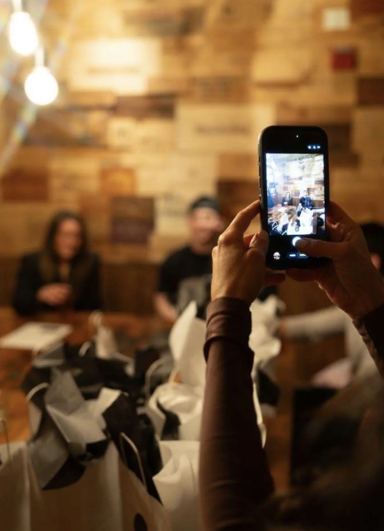 Person taking a photo of two people sitting at a table in a restaurant with a wood-paneled wall in the background.
