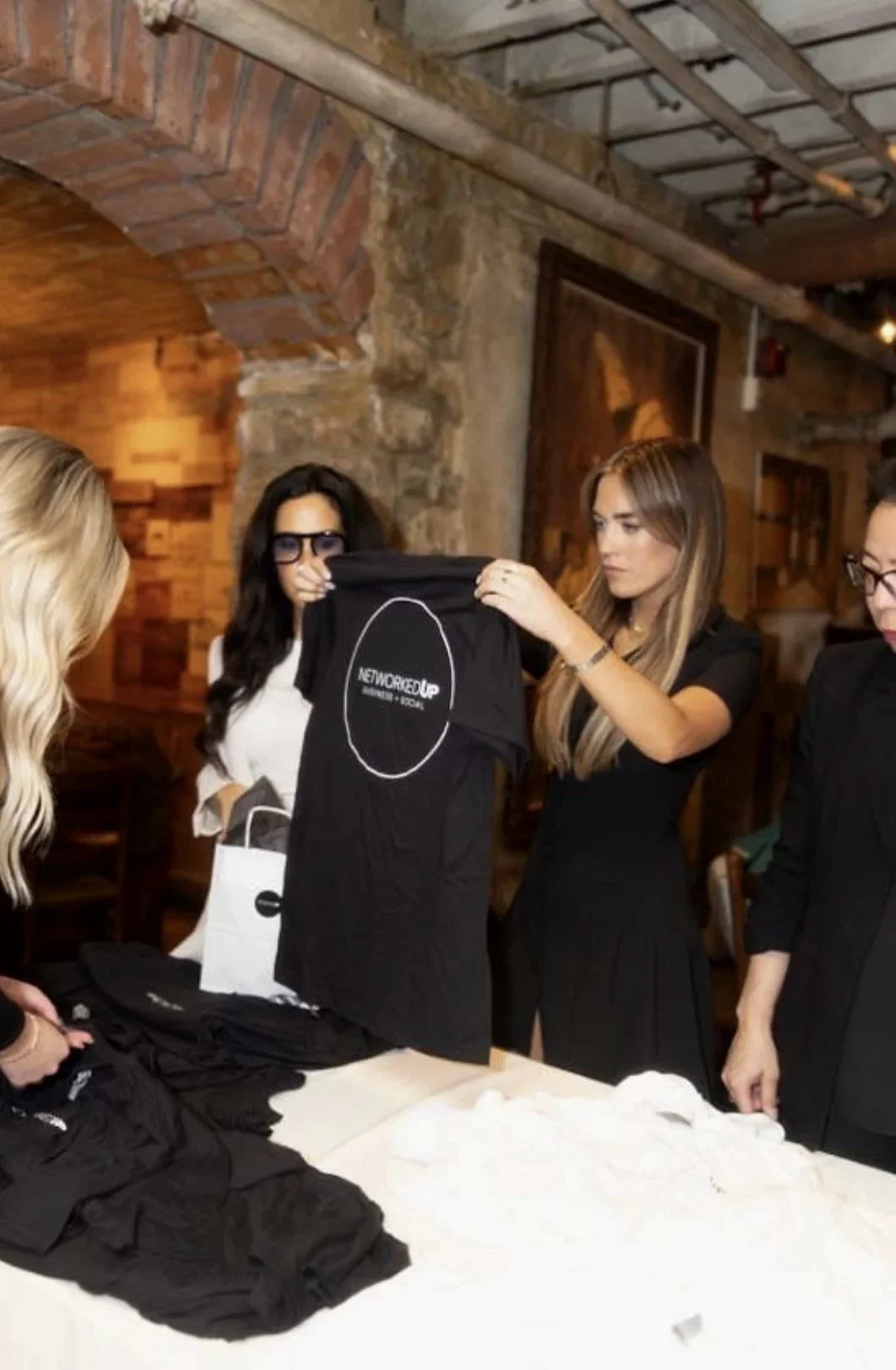 Women at a table displaying black t-shirts, one woman holding up a t-shirt with a white outline of a circle and the words 'NETWORKED UP' printed on it in a restaurant or cafe with brick walls.