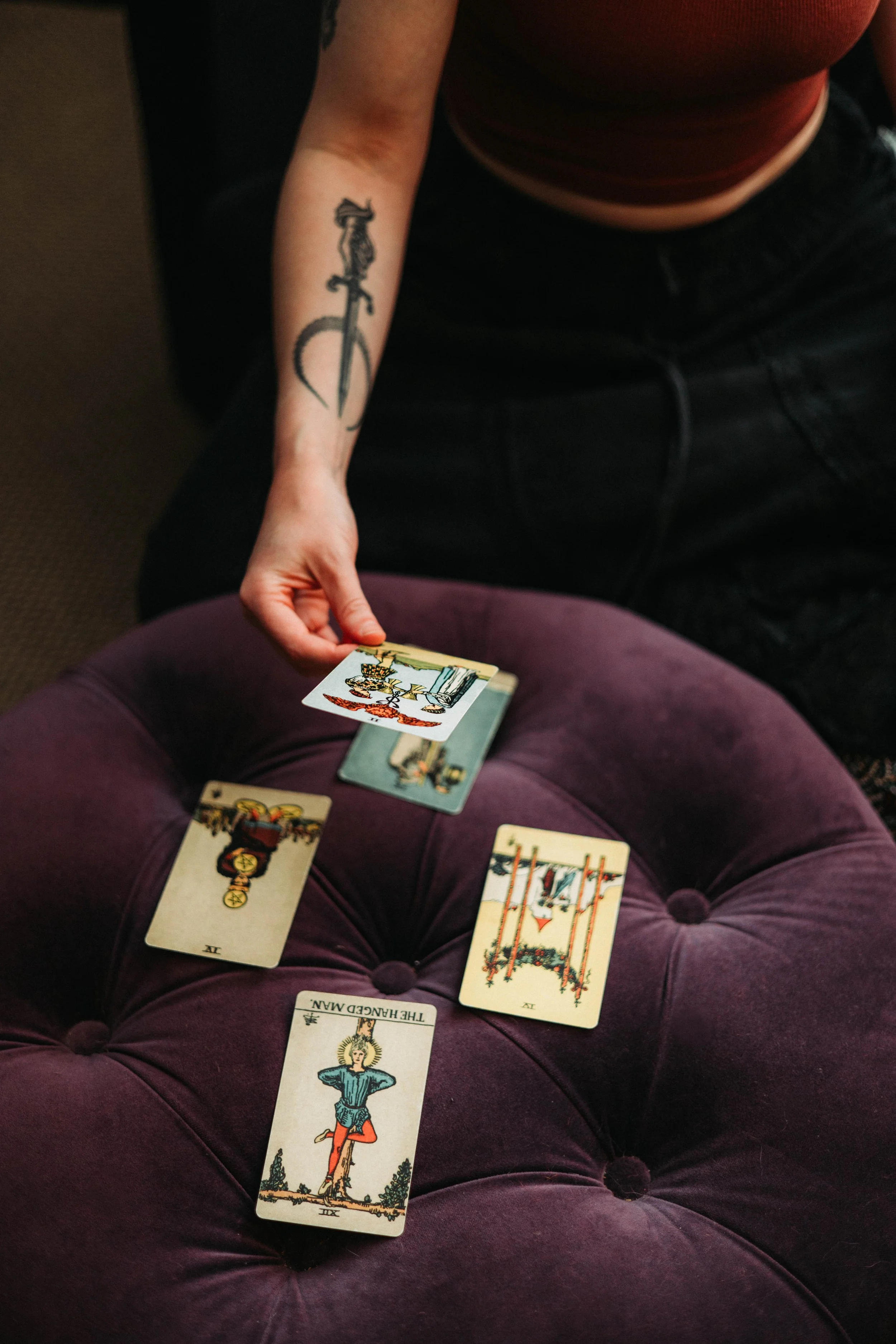 A person with a tattoo on their arm holding a tarot card deck, with several tarot cards spread out on their lap and a few more cards on a purple cushioned surface.