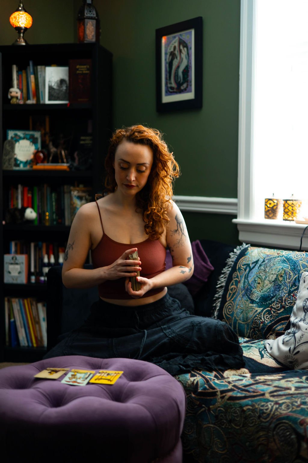 sitting on a patterned couch in a cozy living room. She is holding a deck of tarot cards. The room has a dark bookshelf filled with books and decorative items, with framed artwork on the wall and candles on the window sill. A purple ottoman with tarot cards on top is in front of her.