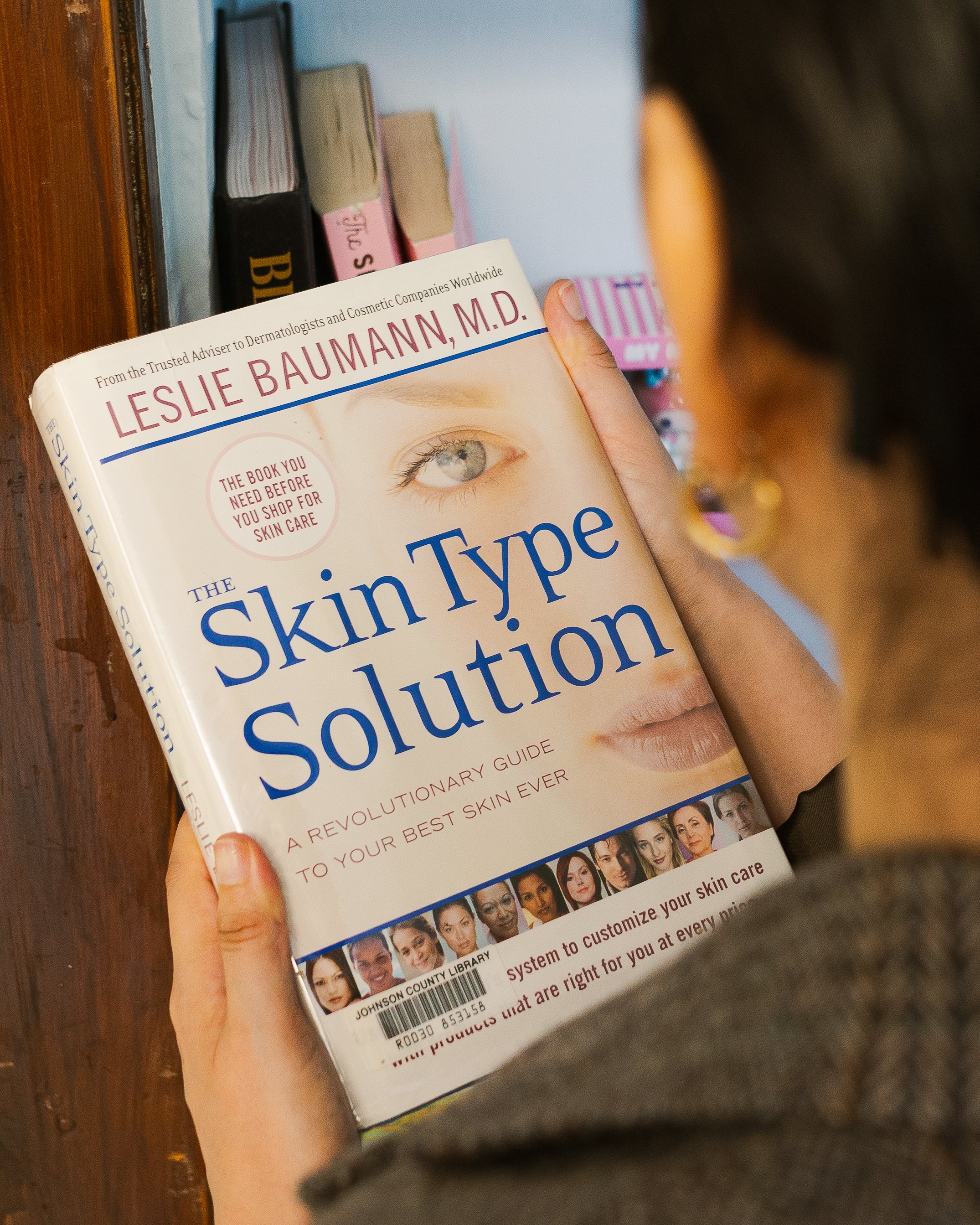 A person holding a book titled 'The Skin Type Solution' by Leslie Baumann, M.D., with a background of other books on a wooden surface.