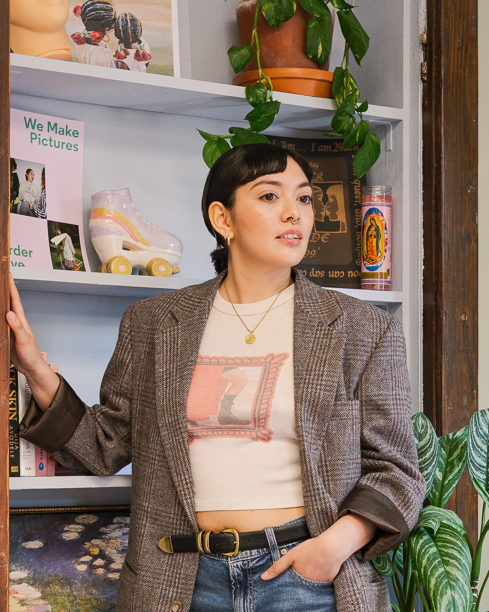 A woman with short dark hair, wearing a beige graphic t-shirt, a gray blazer, and blue jeans, standing indoors in front of a white shelf with plants and artwork, with her left hand on her hip.
