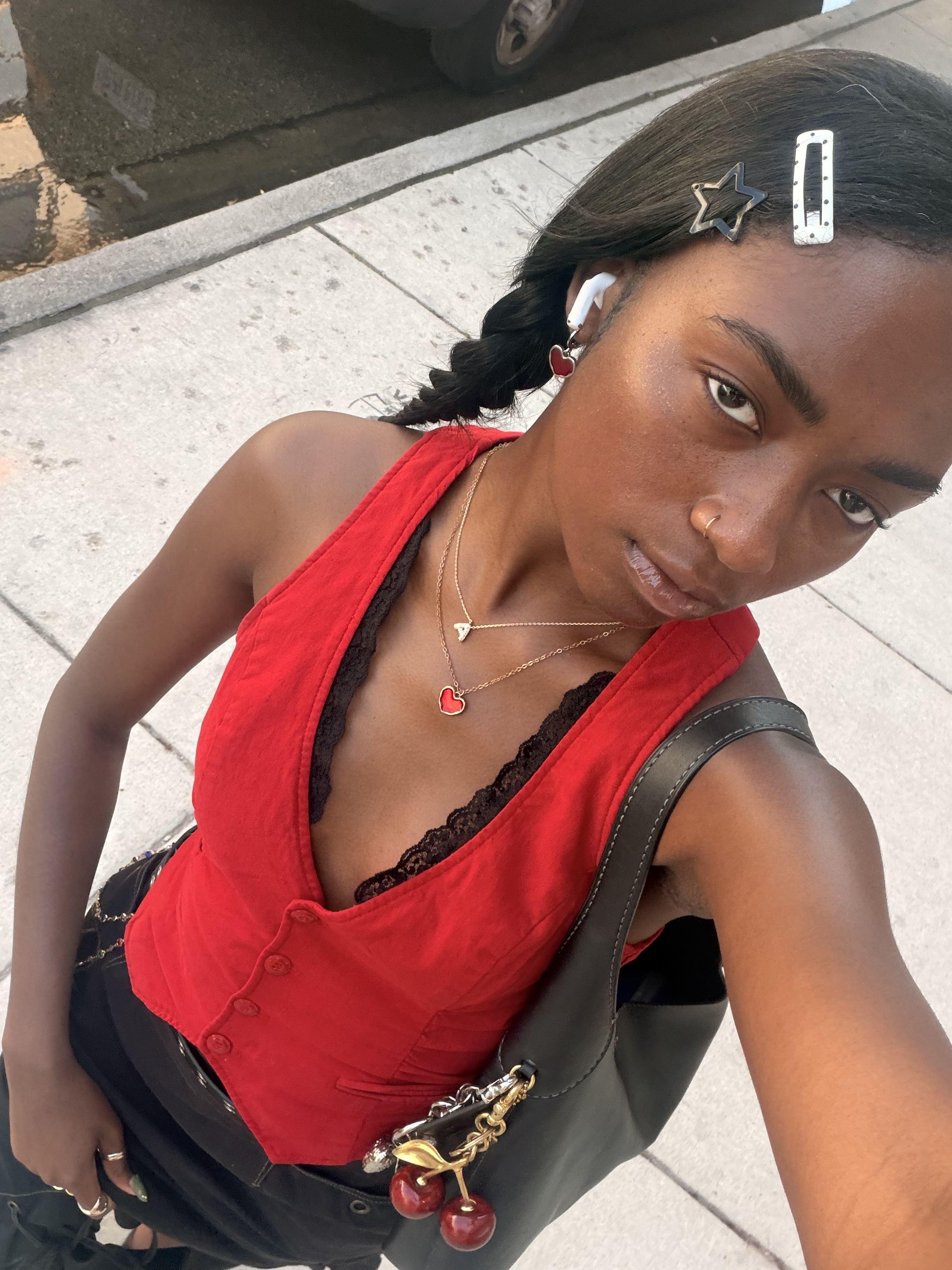 A young woman taking a selfie outdoors, wearing a red sleeveless top with a black lace trim, black pants, and accessories including layered necklaces, earrings, and hair clips with star and rectangular shapes. She has a nose piercing and is carrying a black purse with a chain strap and red cherry keychain. Part of a parking lot and sidewalk are visible in the background.