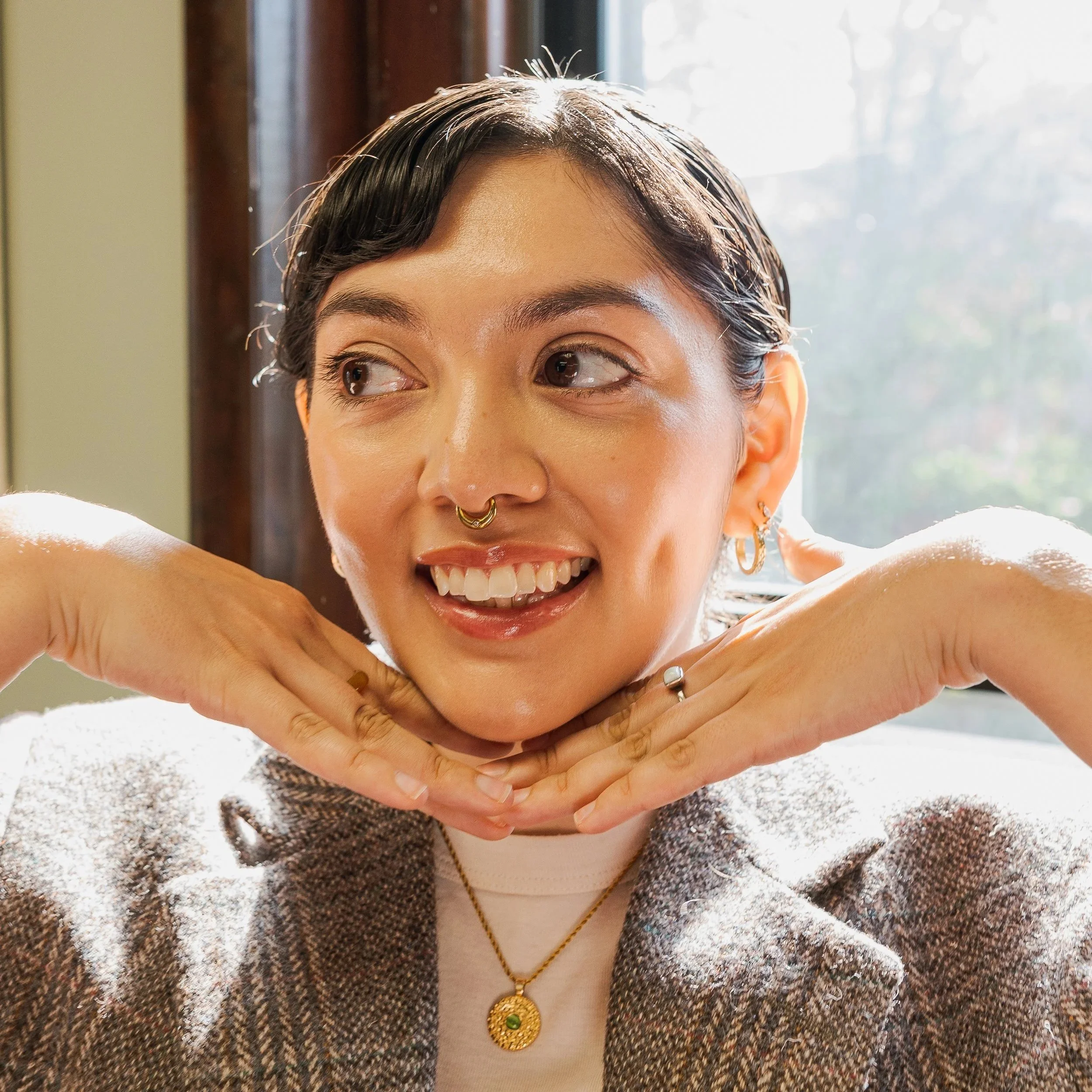 Close-up of a woman with short dark hair smiling, wearing a gold necklace with a round pendant, a bracelet, rings, earrings, and a brown sparkly jacket, with sunlight coming through a window behind her.