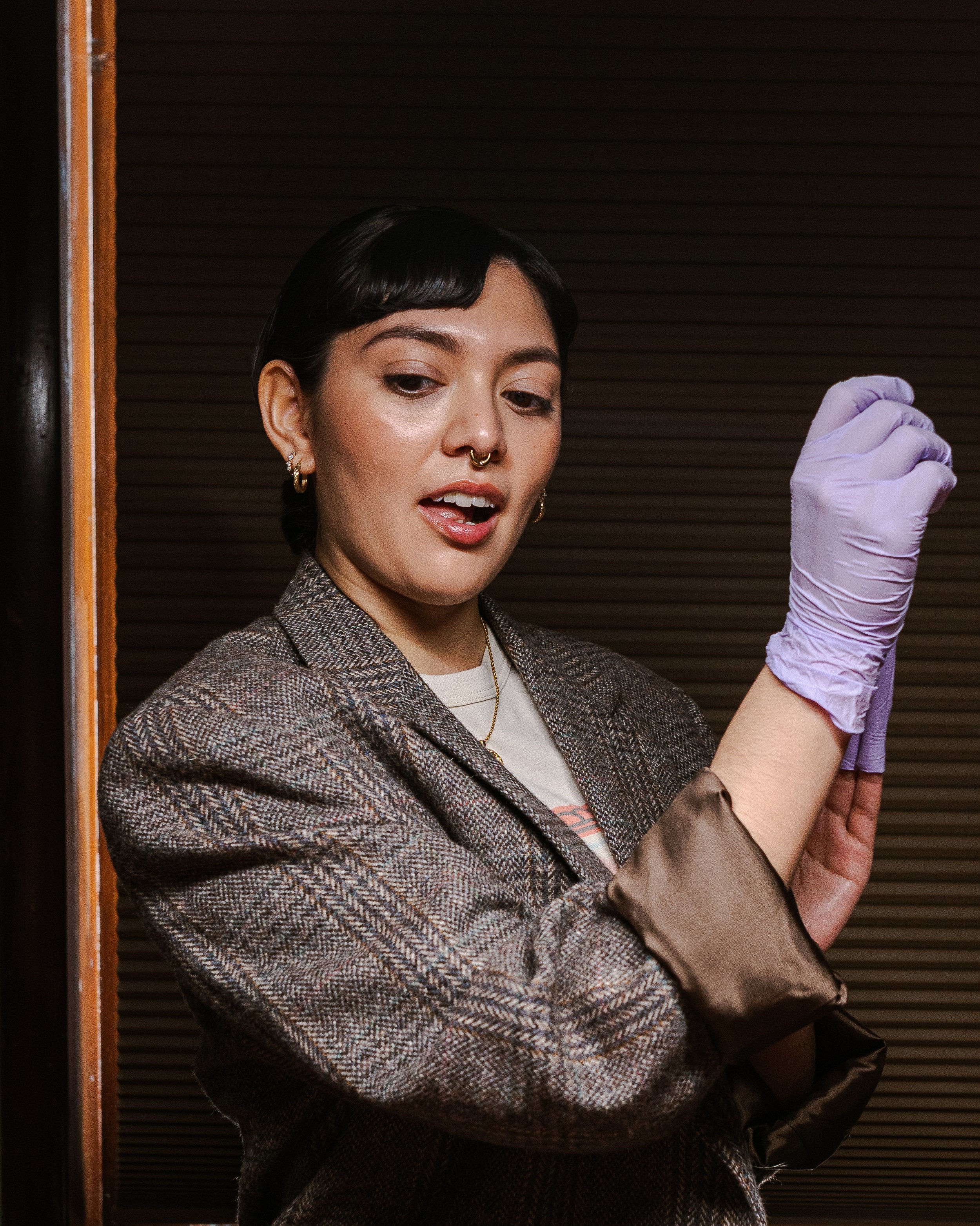 A woman with black hair, wearing earrings, a septum piercing, and a checked blazer, is putting on a purple glove in front of a brown wooden background.