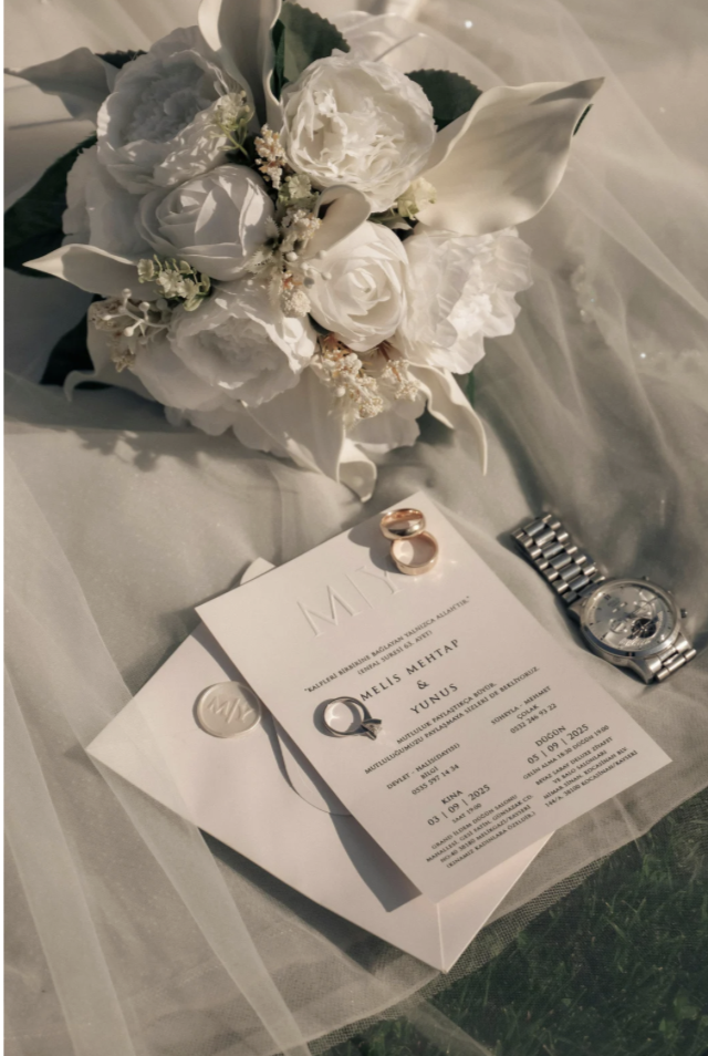 Wedding invitation card, bouquet of white flowers, gold rings, silver watch, and a white envelope on a wedding table.