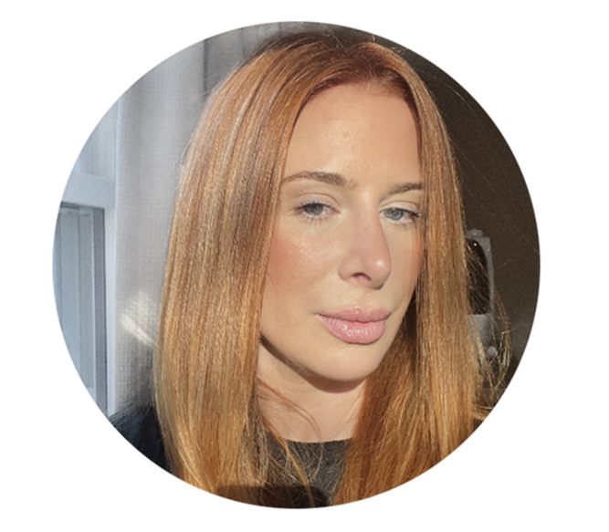 Close-up of a woman with straight red hair, light skin, and blue eyes, looking slightly to the side with a neutral expression, standing near a window with sunlight illuminating her face.