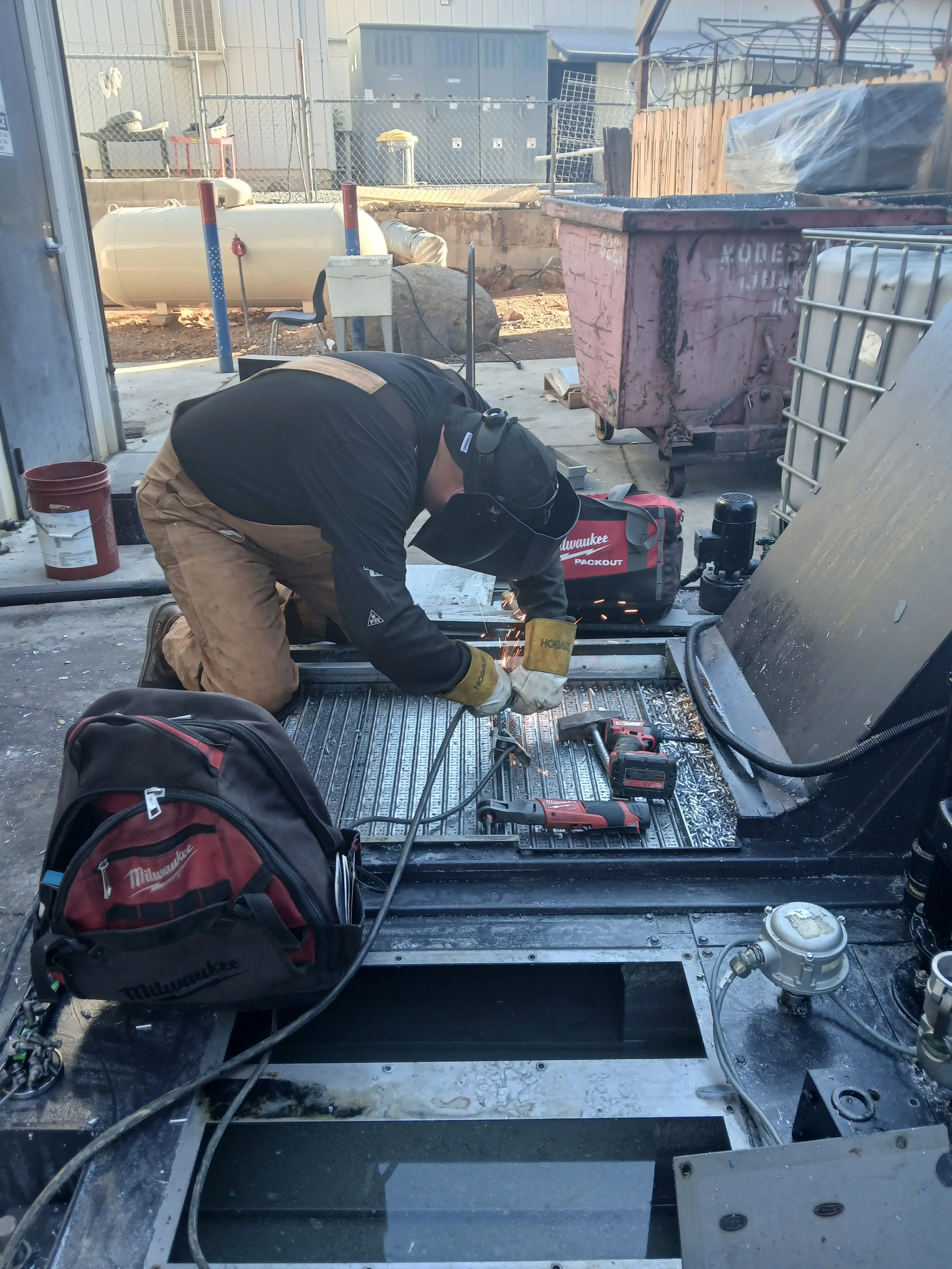 A worker welding metal on a construction site. The worker is wearing protective gear including a welding helmet, gloves, and a black jacket with tan pants. Tools and equipment are visible around him, including a red Milwaukee backpack, a cordless drill, and a red toolbox.