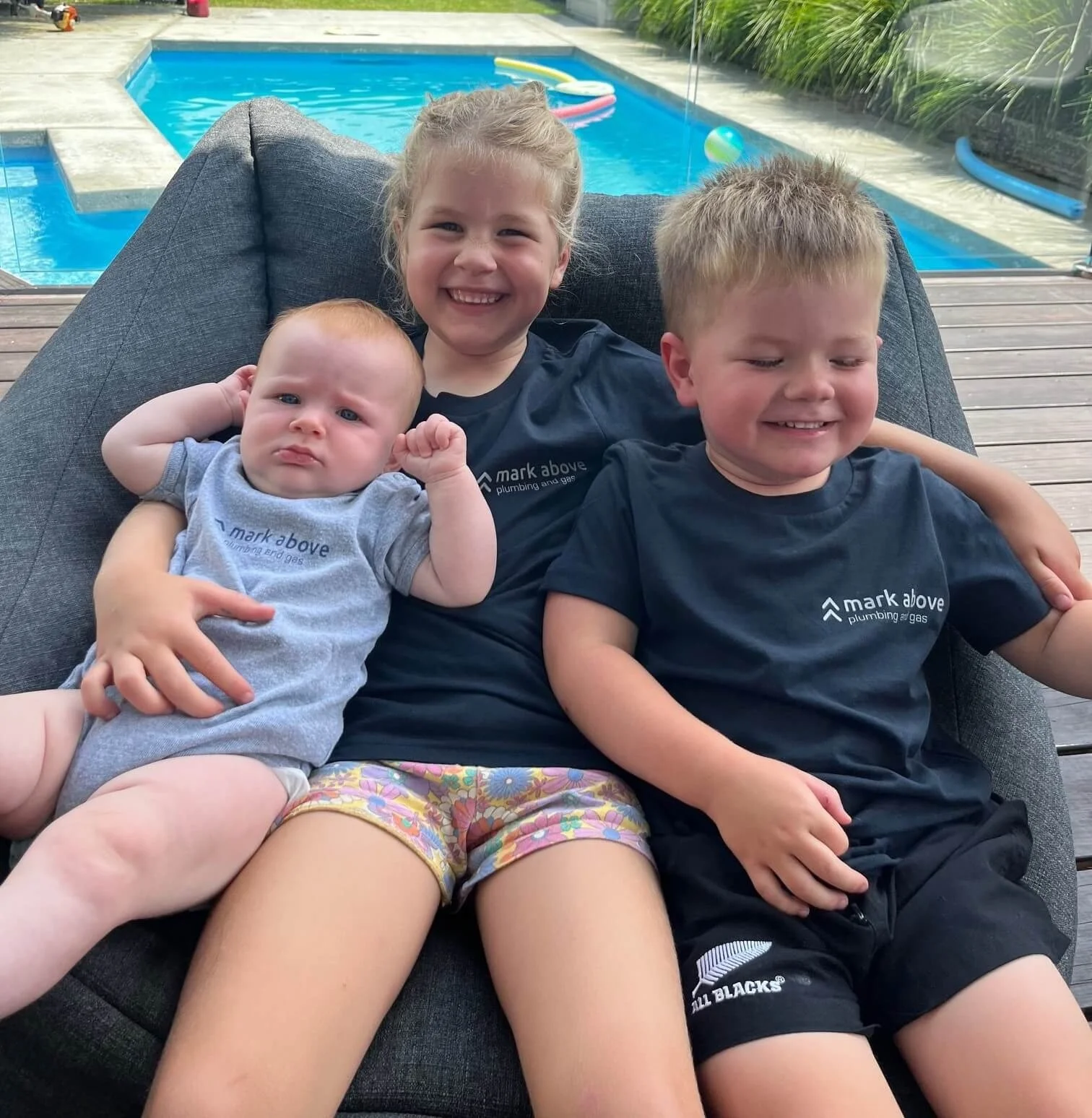 Three children sitting outdoors on a black couch with a swimming pool and lush greenery in the background. The youngest, a baby, is on the left, lying on a woman's lap, wearing a gray shirt and colorful shorts. The girl in the middle, smiling widely, wears a black shirt with a logo. The boy on the right, also smiling, wears a matching black shirt and black shorts, with his arm around the girl.