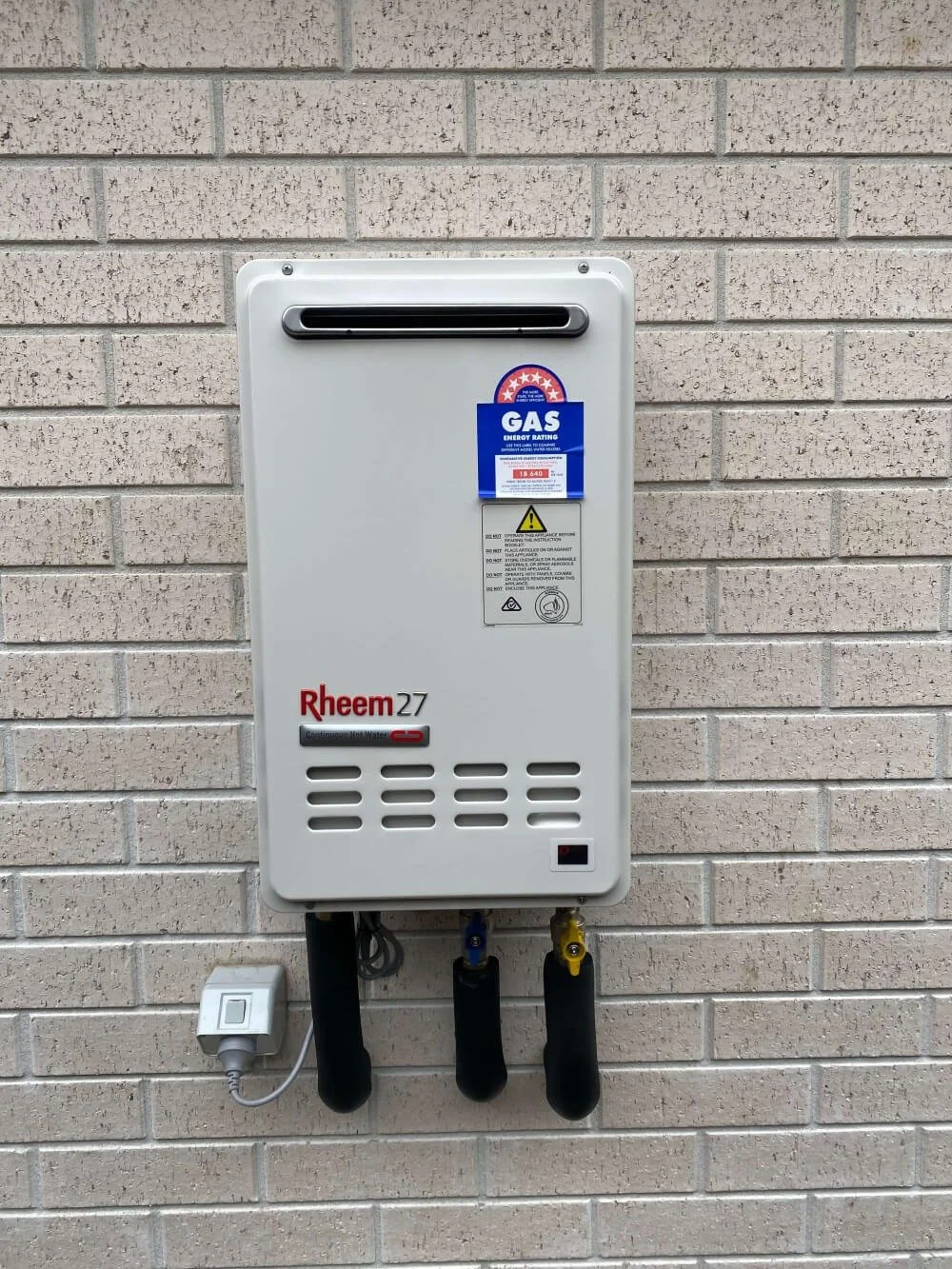 A Rheem brand gas water heater mounted on a beige brick wall with a power cord plugged in and three pipes connected at the bottom. There are labels and warning signs on it, including a blue sticker indicating gas energy rating.