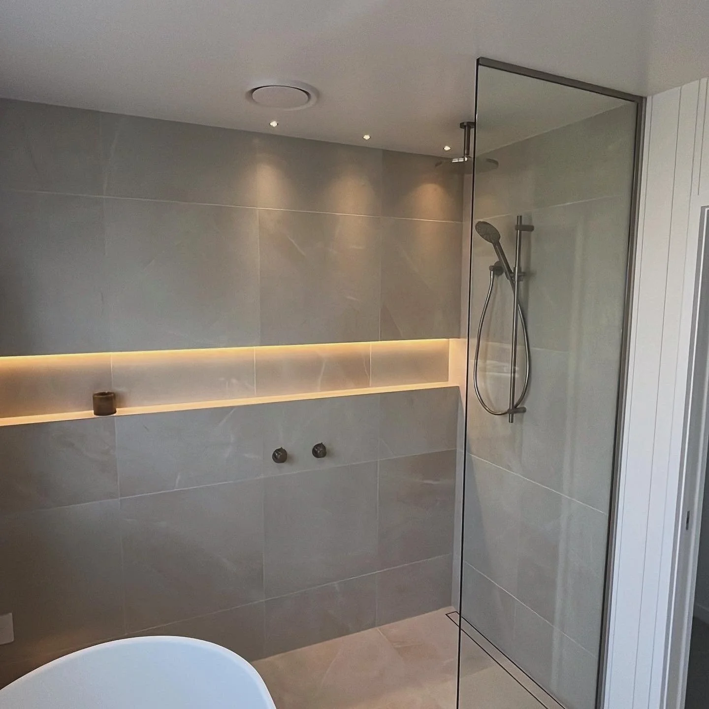 Modern bathroom with a walk-in shower enclosed by a glass panel, grey tiled walls, and built-in shelf with warm lighting.