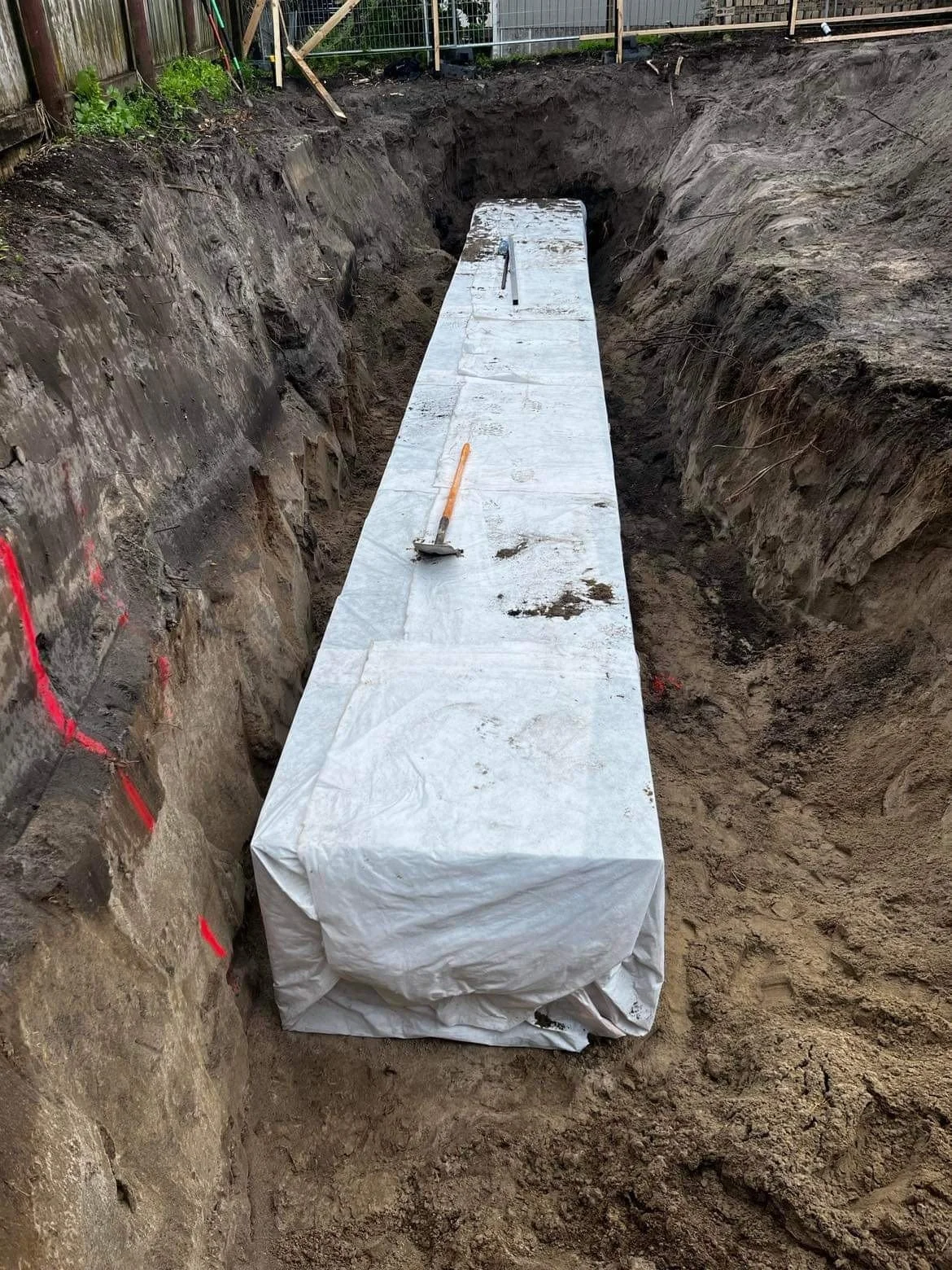 A construction site showing a large wrapped rectangular object placed in a deep trench with rocky and dirt walls, and a groundwater portion at the bottom.