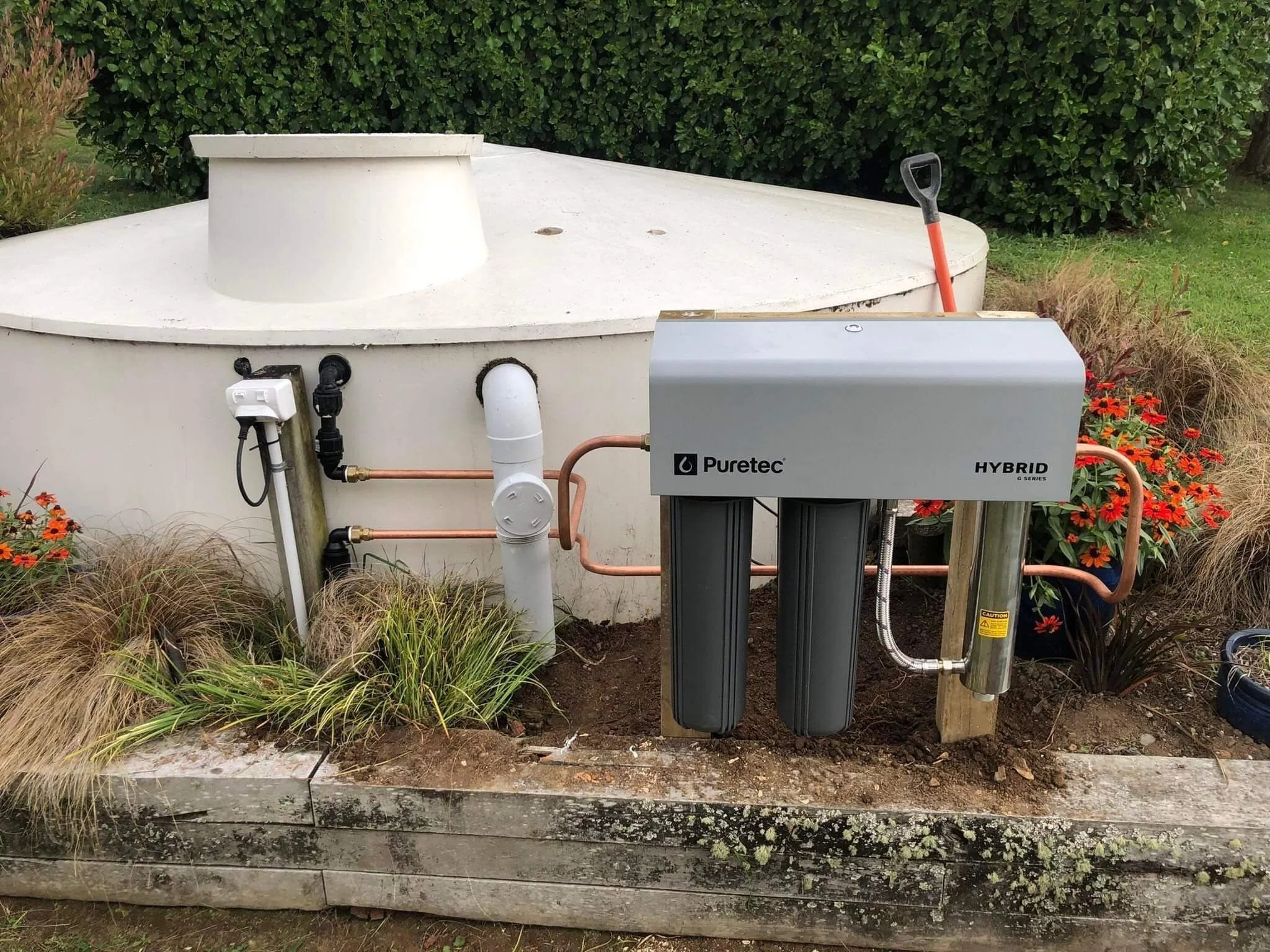 An outdoor scene with a white water heater or filtration system labeled "Puretec HORSE series" connected to pipes, with flower plants and greenery surrounding it.