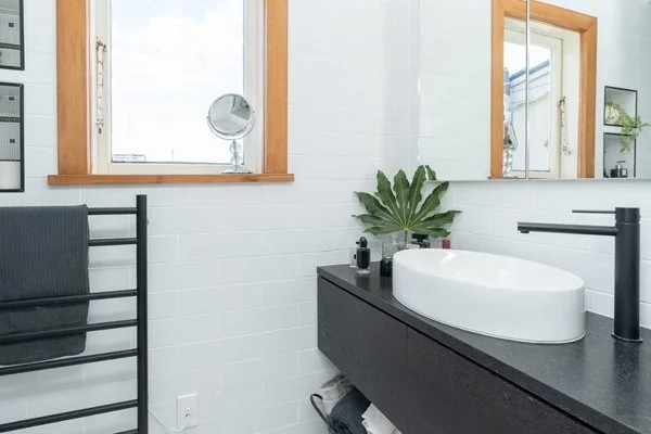 Modern bathroom with white tiled walls, a black countertop with a white oval sink, a black faucet, a large mirror, a small vanity mirror, green plant, black towel rack, and windows with wooden frames.
