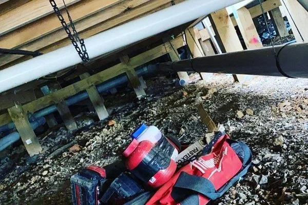 Underneath a building or deck, showing pipes, a chain, and a red and black tool bag on dirt and gravel.