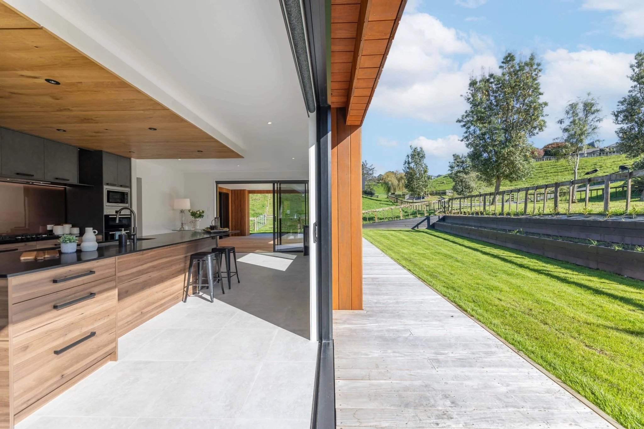 View from inside a modern kitchen looking outward to a wooden deck and a lush, green yard with trees under a partly cloudy sky.