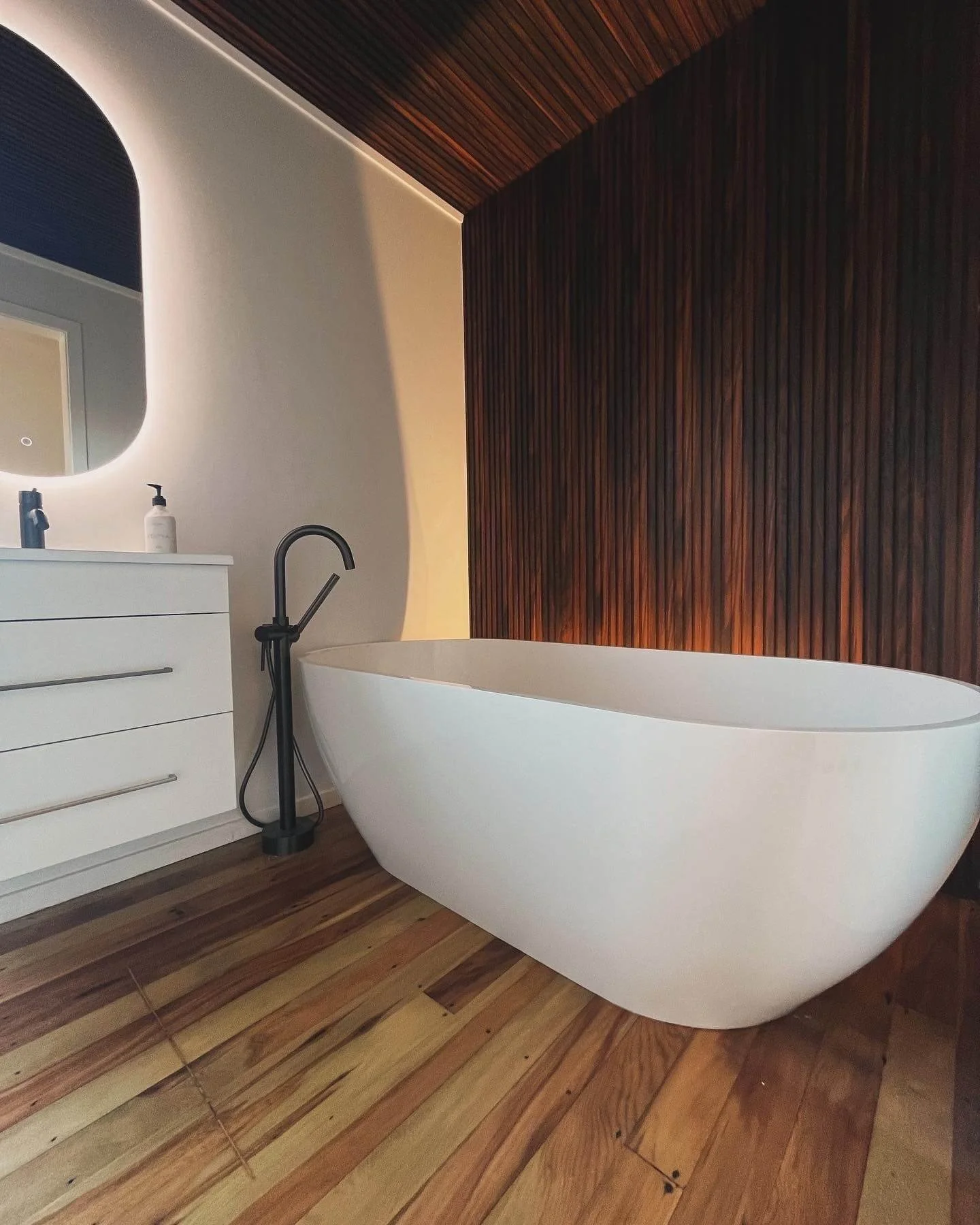 Modern bathroom with a white freestanding bathtub, wooden flooring, a white vanity with a mirror, and a wooden accent wall behind the bathtub.
