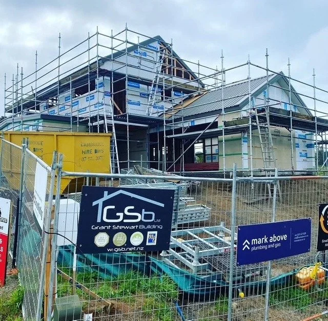 Construction site of a house with scaffolding around it, showing a partially built roof and walls, with construction signs and a yellow skip bin in the foreground.